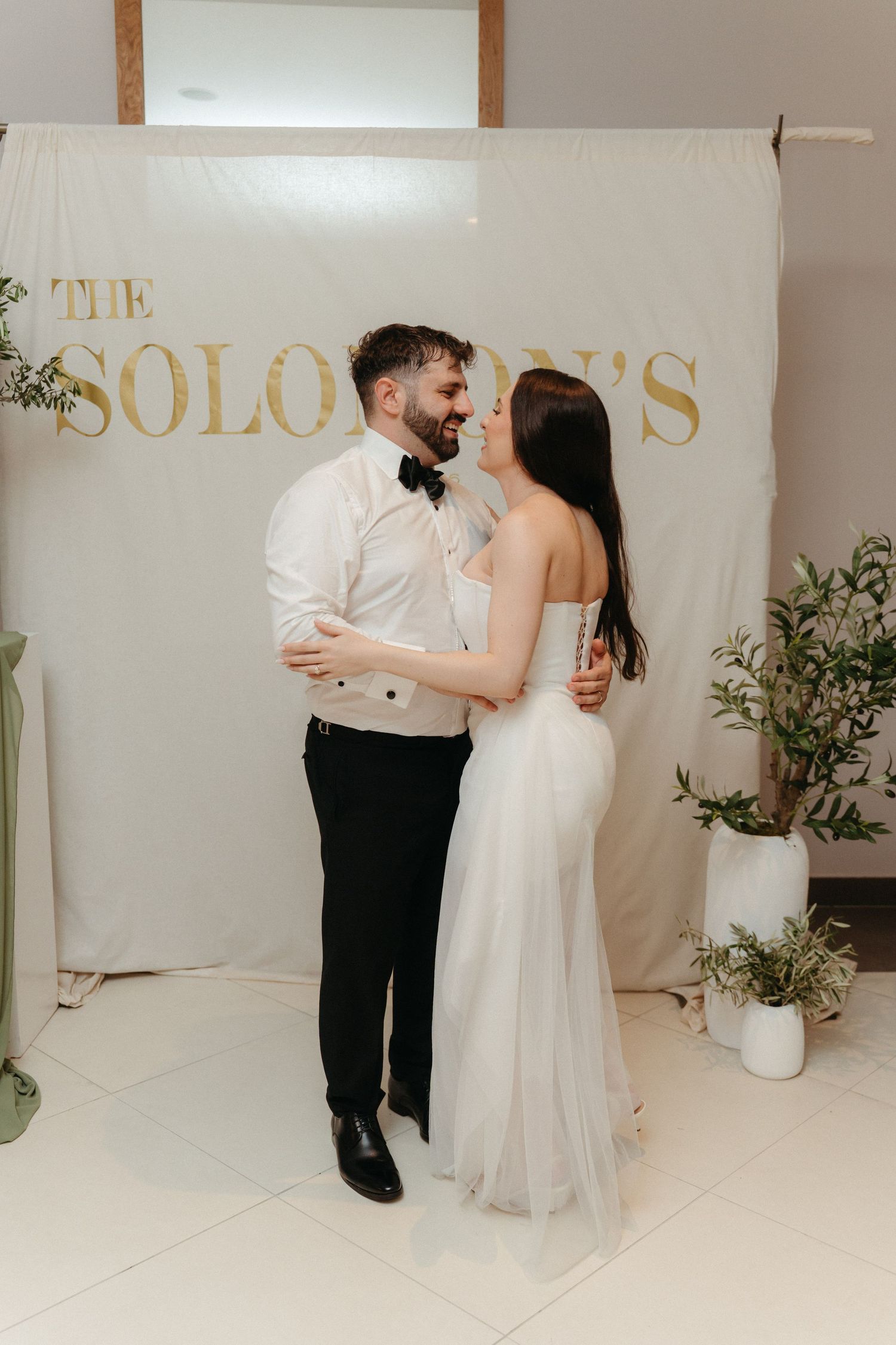 Bride and groom share a romantic moment in front of The Solomons wedding backdrop, surrounded by greenery.