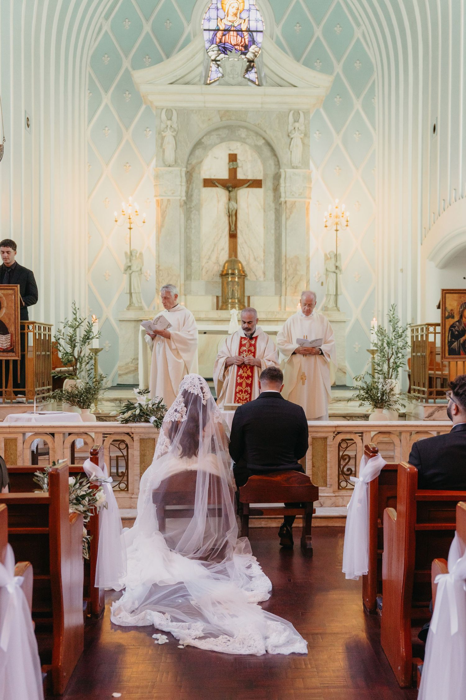 Bride and groom kneel before priest at ornate church altar during traditional Catholic wedding ceremony.