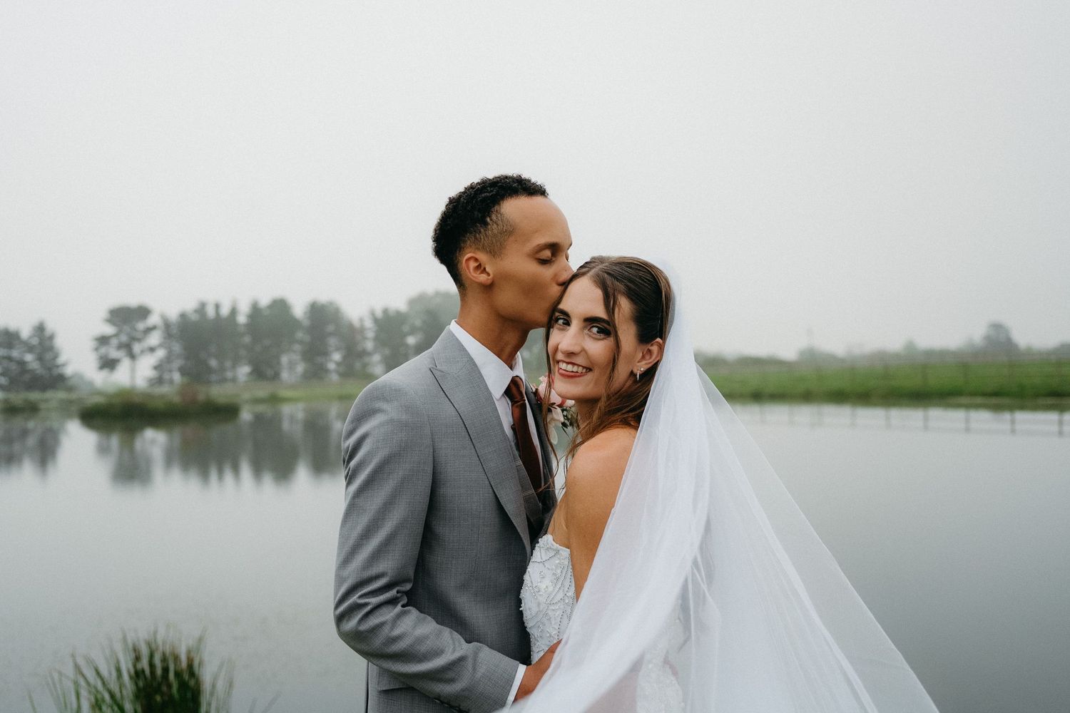 A couple shares a romantic kiss on their wedding day beside a misty lake, bride in white gown with veil, groom in gray suit.