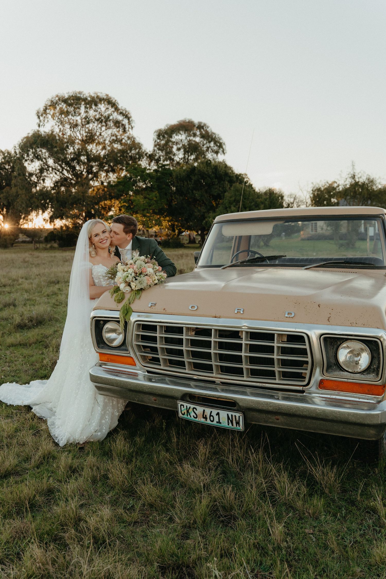 Bride and groom pose beside a vintage Ford pickup truck at golden hour on a rustic farm wedding venue.