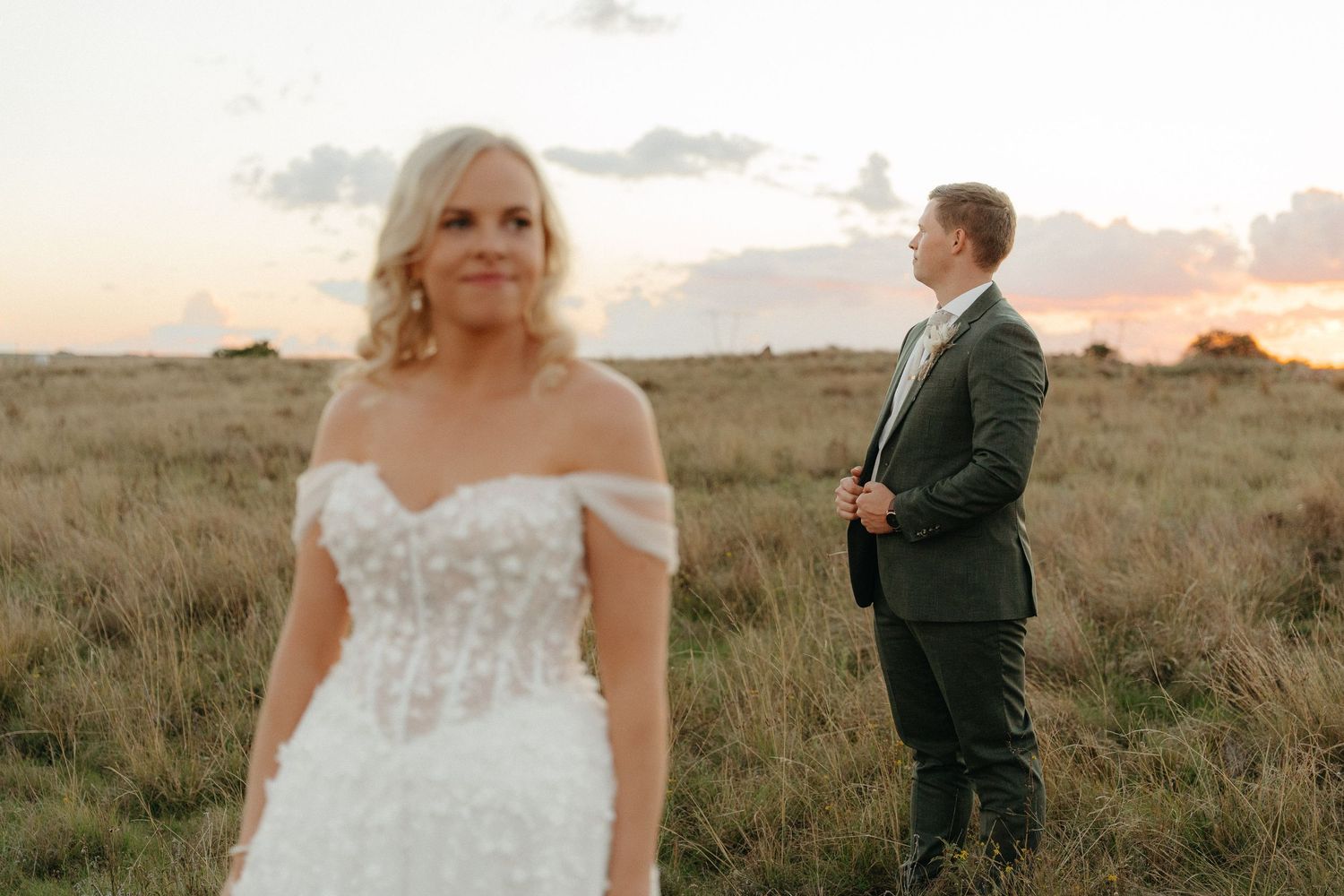 Bride in off-shoulder lace gown stands before groom in dark suit at golden sunset over open grassland fields.