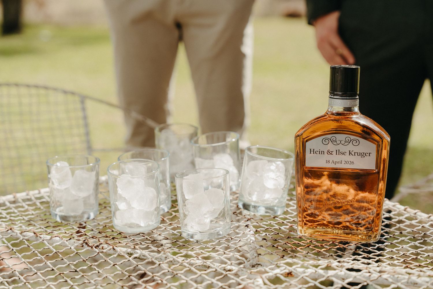 Outdoor whiskey bar setup with Hess & the Keeper bottle, ice-filled glasses on a metal mesh table.
