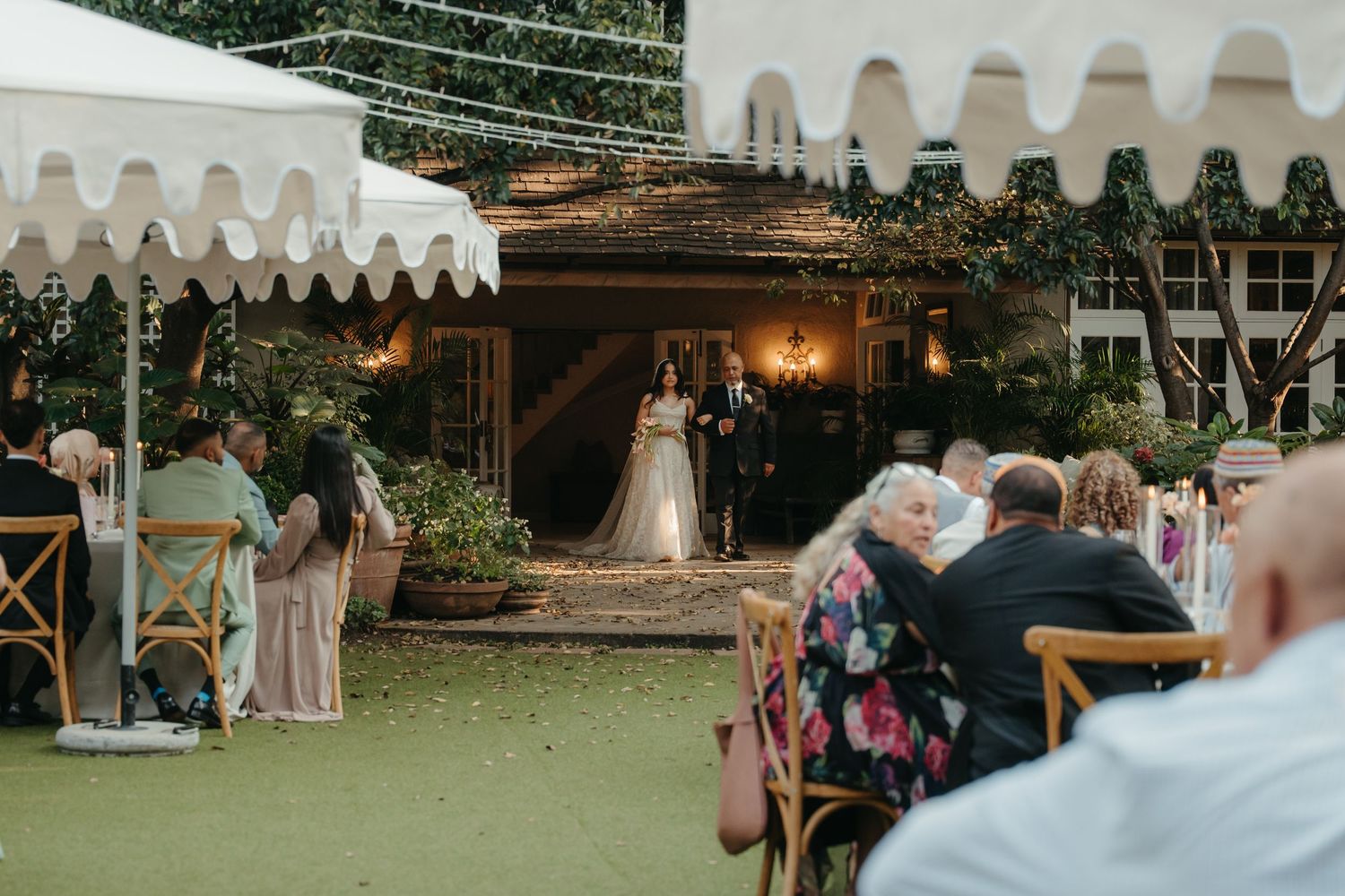 Outdoor wedding ceremony with guests seated on wooden chairs facing couple at altar under string lights and white tent.