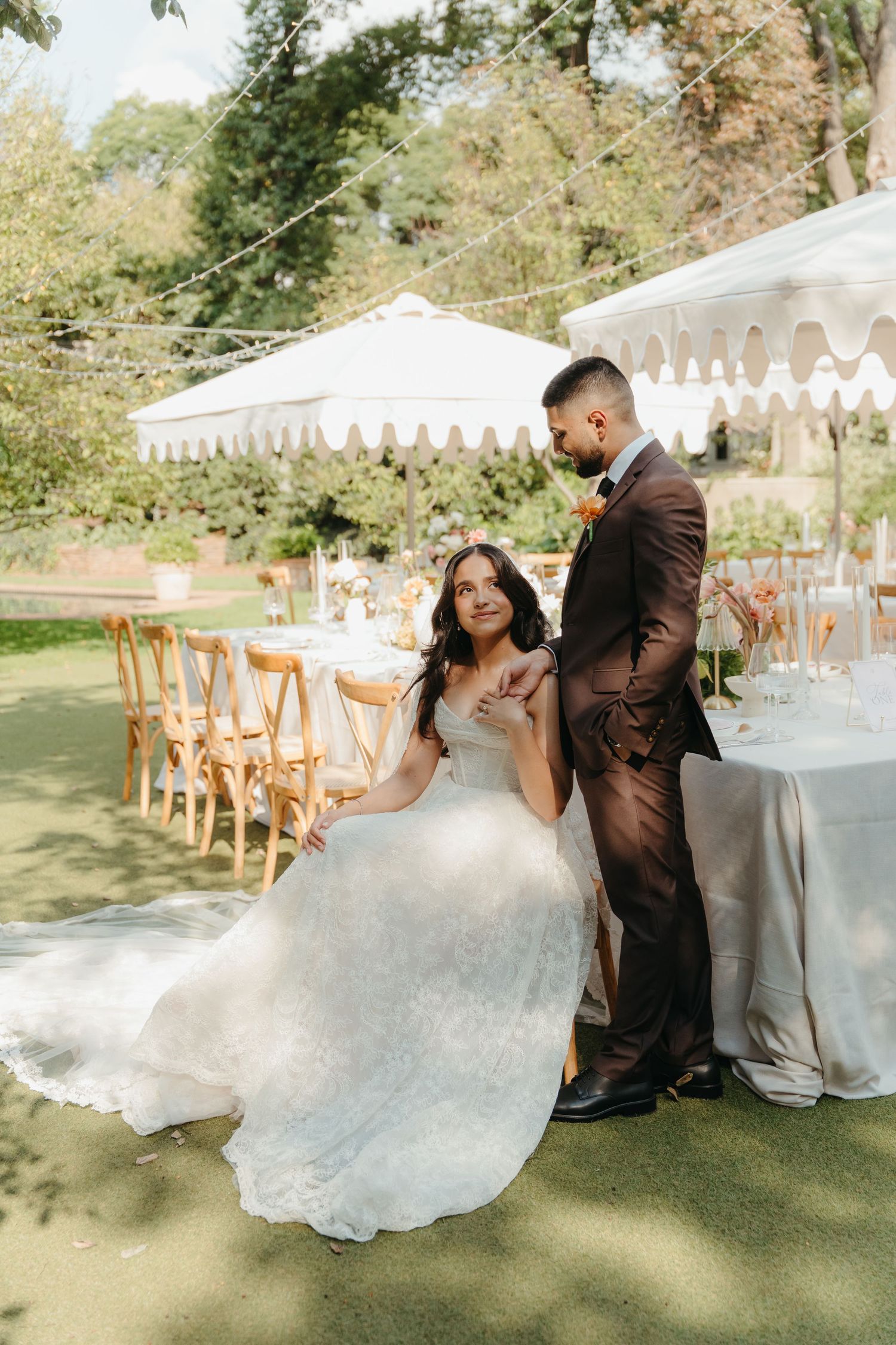Bride in white lace gown sits beside groom in brown suit at elegant outdoor wedding reception under white tent.