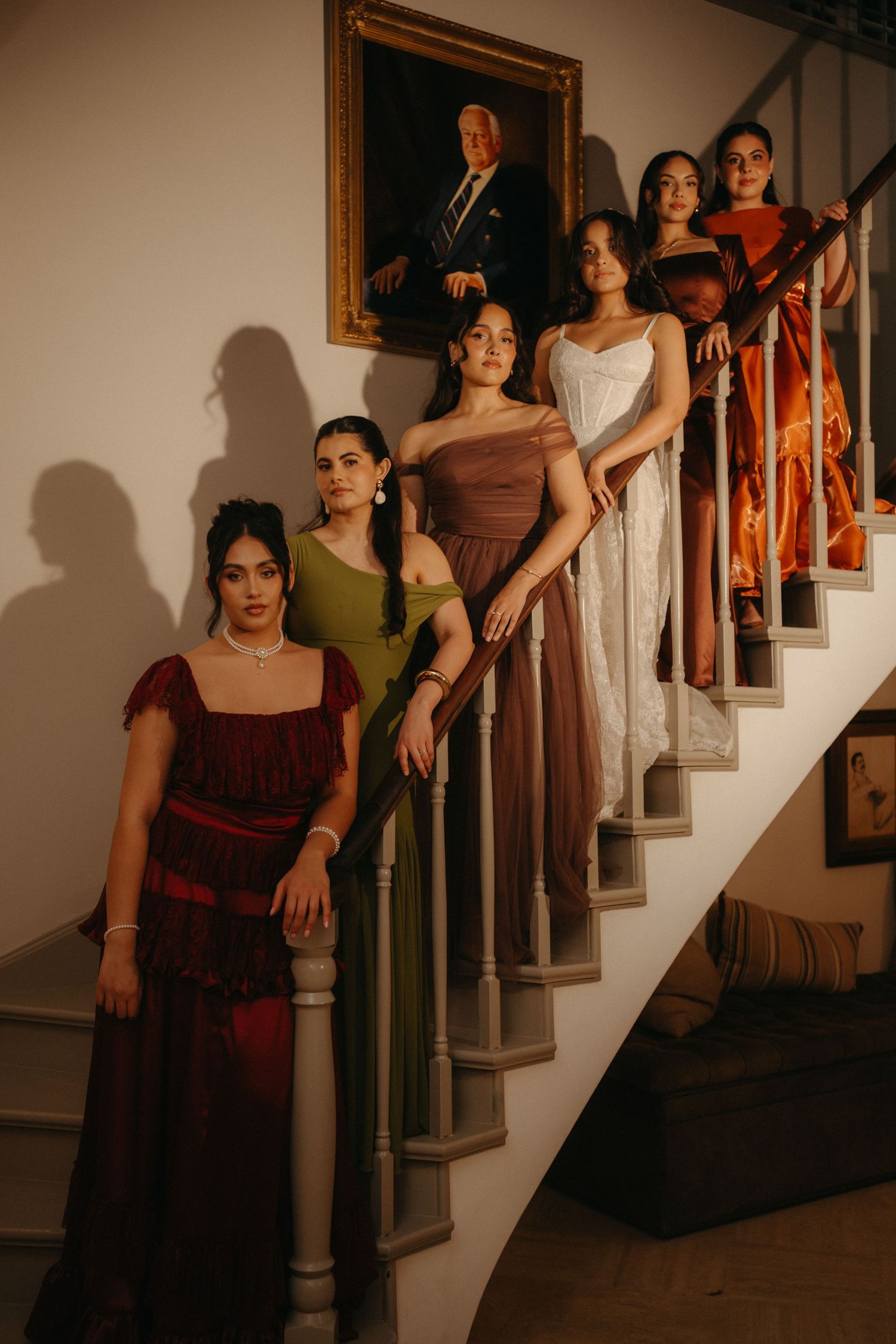 Six women in elegant colorful gowns pose gracefully on a staircase beneath a framed portrait in a warmly lit interior.