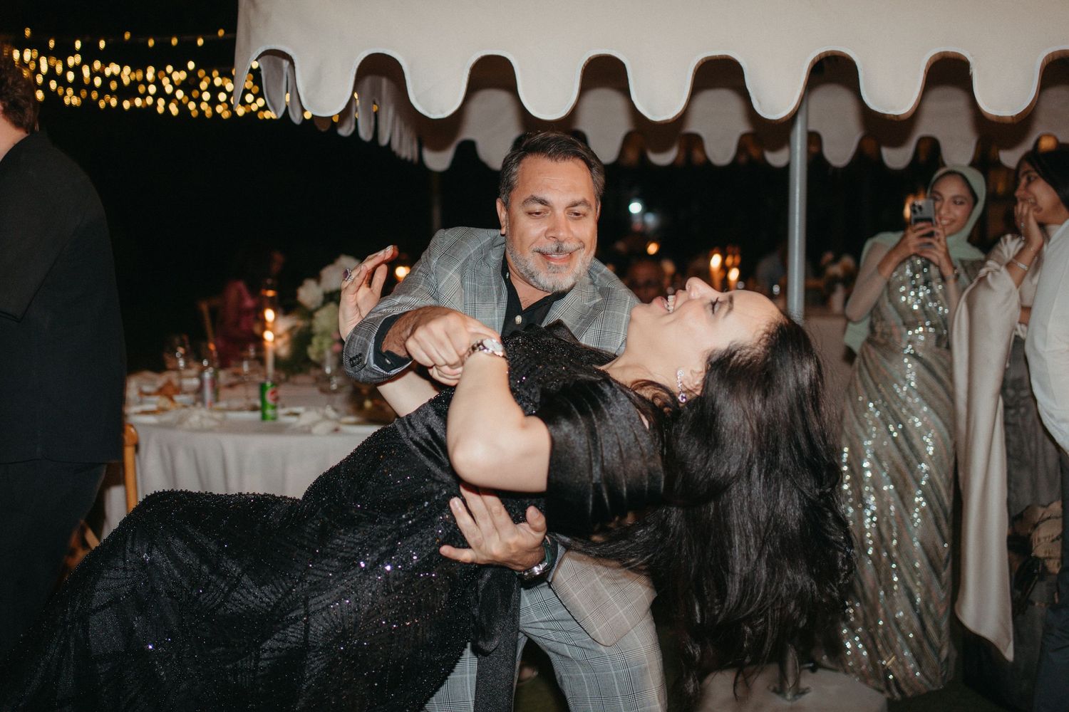 Couple dancing at an elegant outdoor wedding reception under a tent with candlelit tables and guests in background.