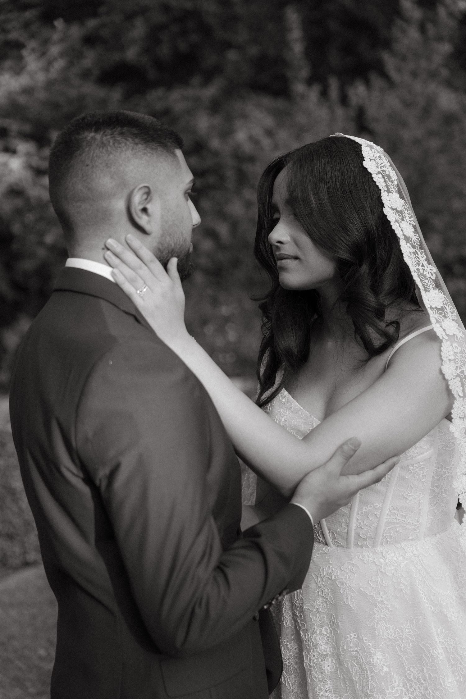 Black and white wedding photo of bride in lace veil tenderly touching groom's face while gazing into each other's eyes outdoors.