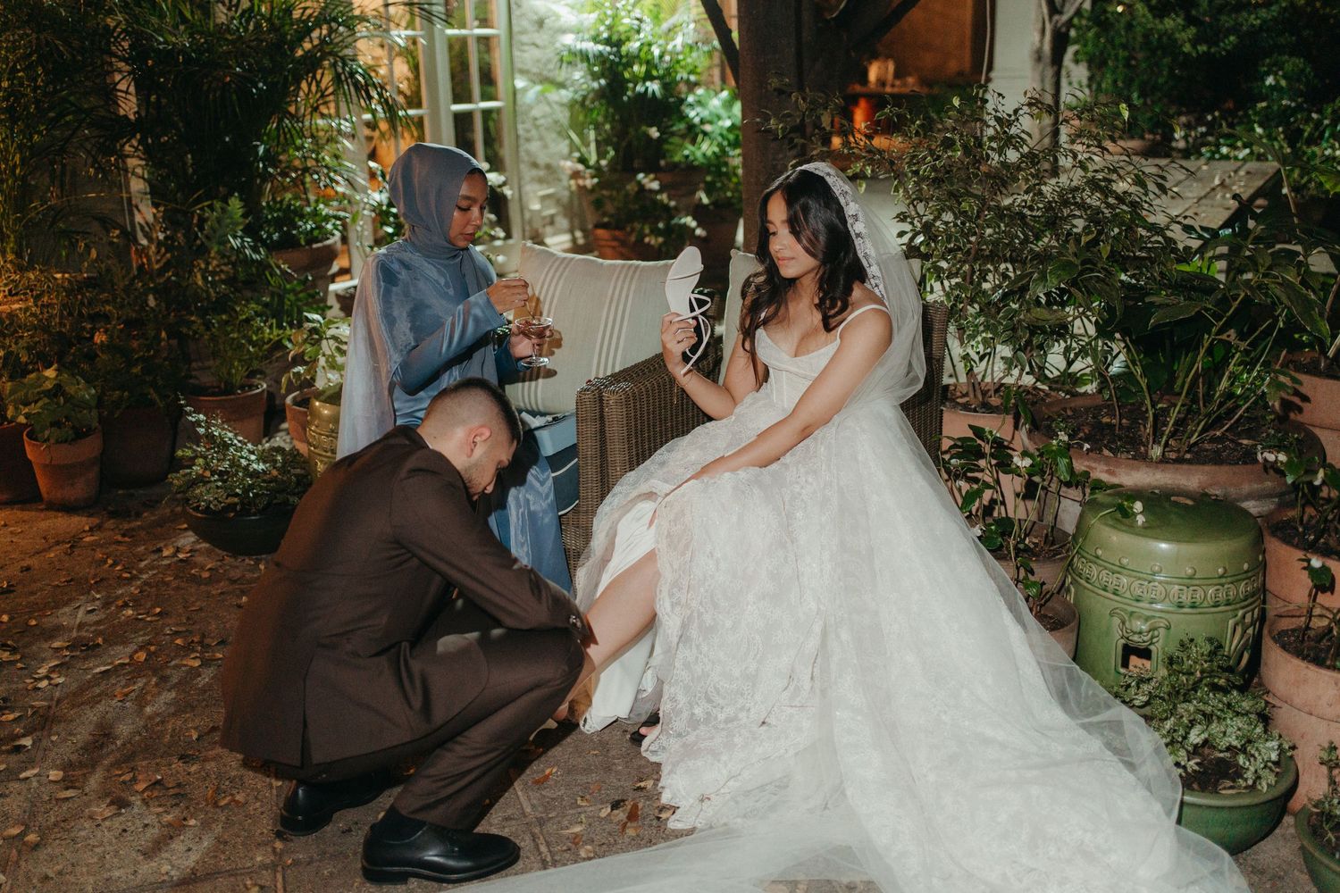 Groom kneeling to adjust bride's wedding dress while guest holds champagne in lush garden setting.