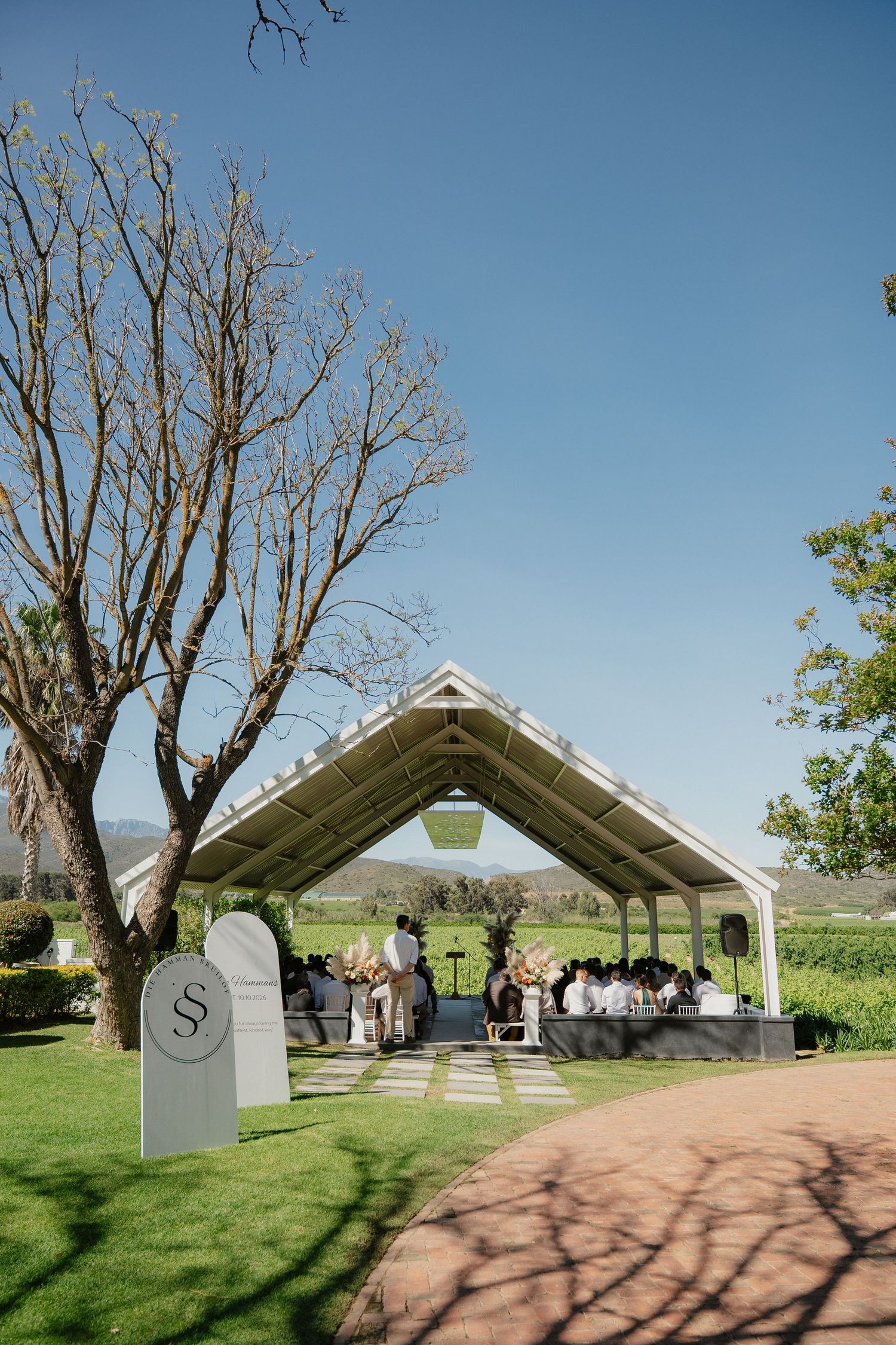Elegant outdoor wedding ceremony under a white pavilion surrounded by lush green gardens and vineyard views.