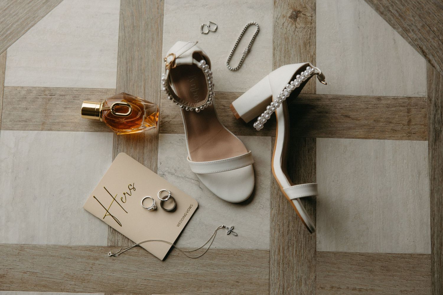 White bridal heels with pearl ankle straps displayed alongside gold rings, perfume bottle, and wedding jewelry on wooden floor.