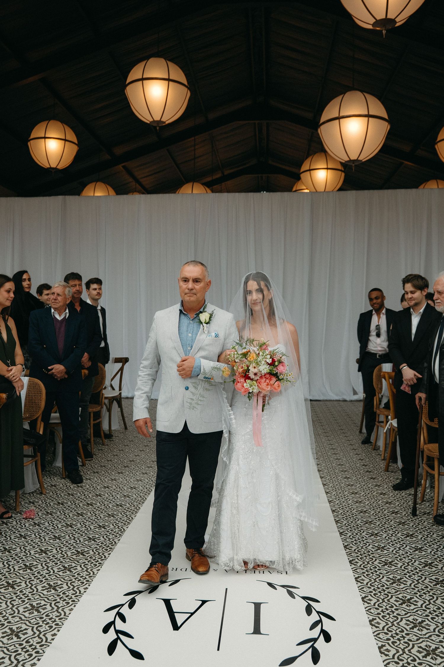 Bride walks down aisle with escort at elegant indoor wedding ceremony with globe pendant lights and monogram aisle runner.