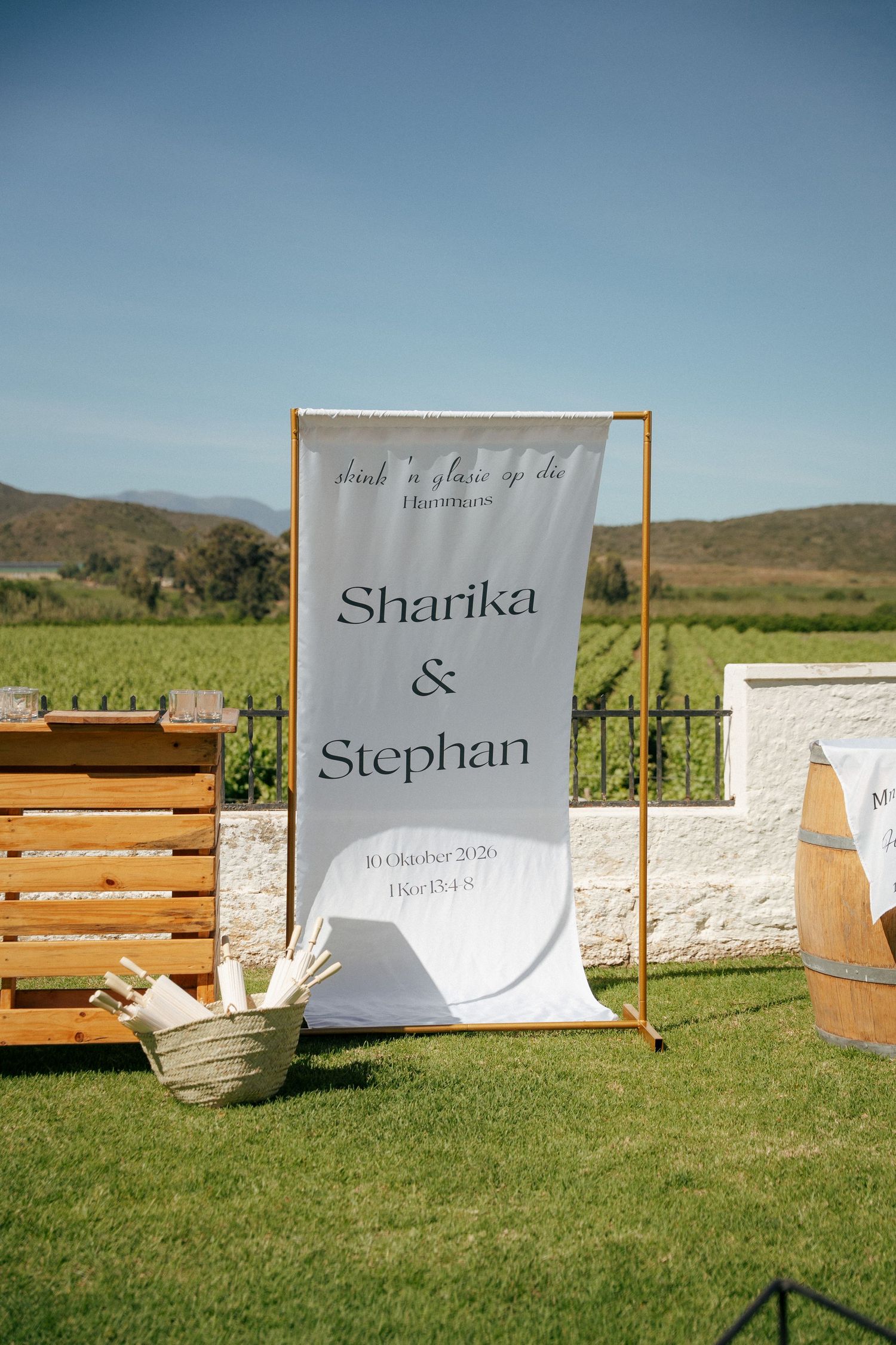Wedding welcome banner for Sharika & Stephan on a gold frame beside a wooden bar and wine barrel, set on a lawn with Cape vineyards behind.