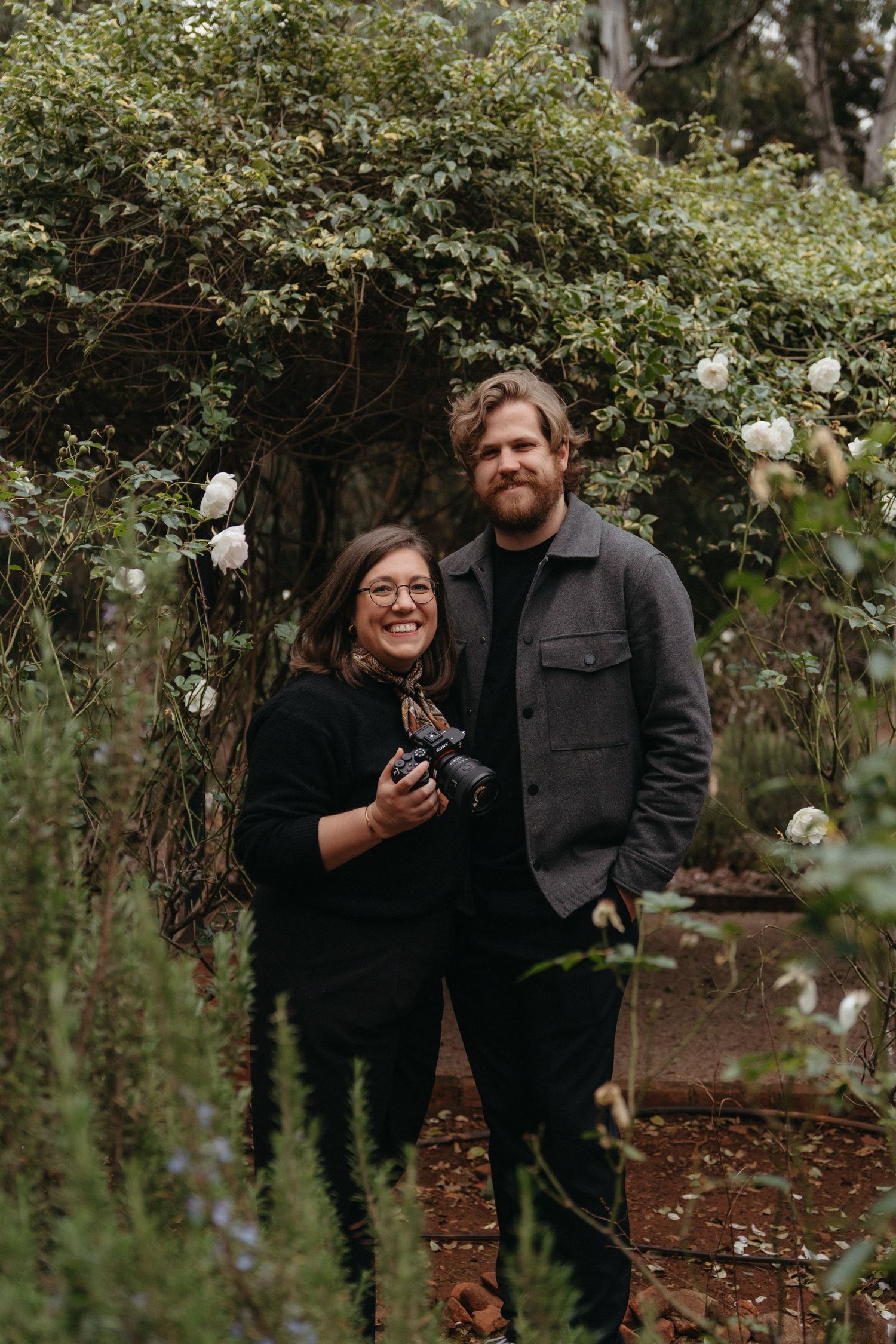 Smiling couple standing in a lush garden with white roses, woman holding a camera, both dressed in black.