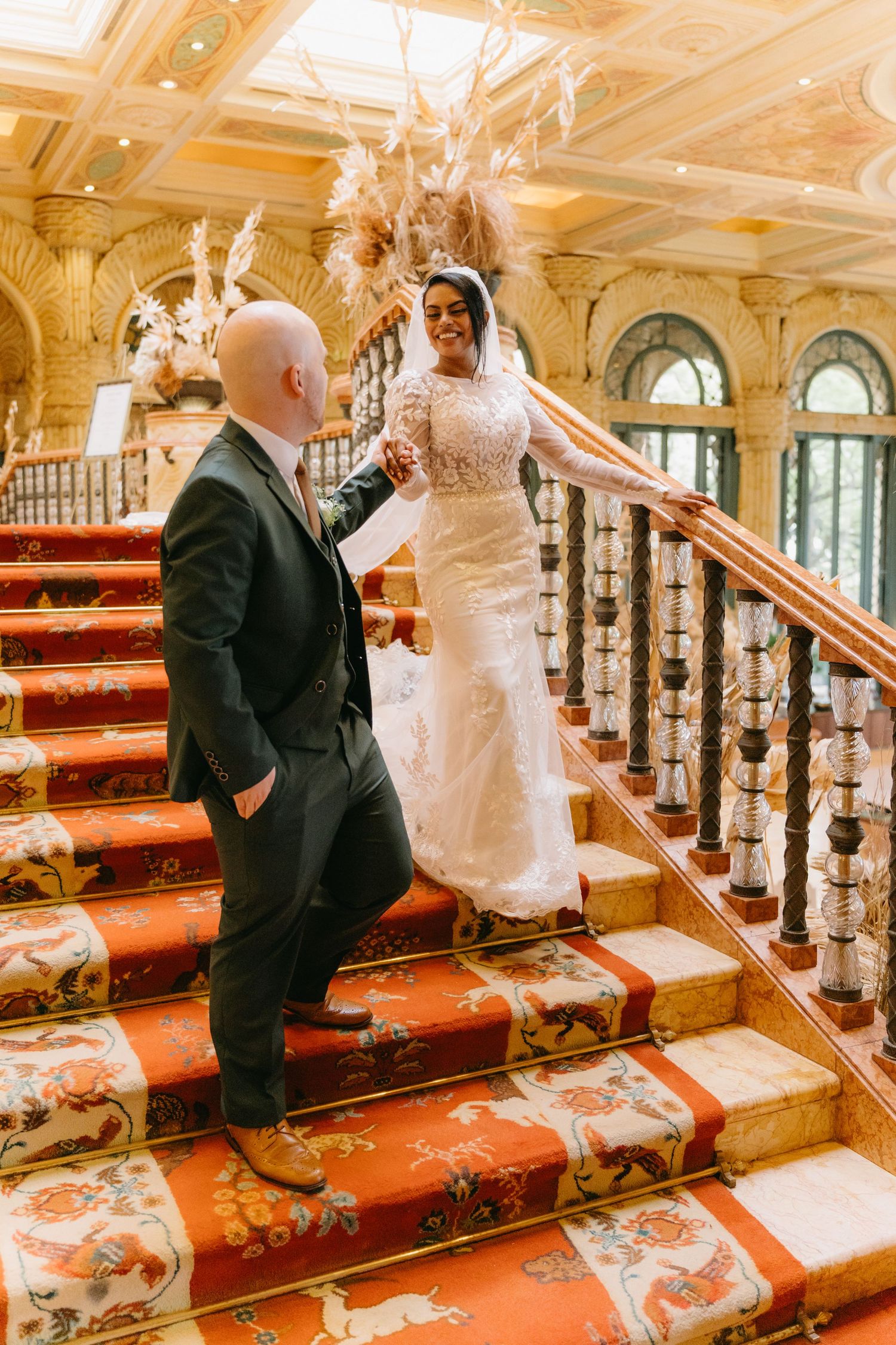 Wedding moment captured on grand carpeted staircase with ornate gold ceiling and decorative railings.