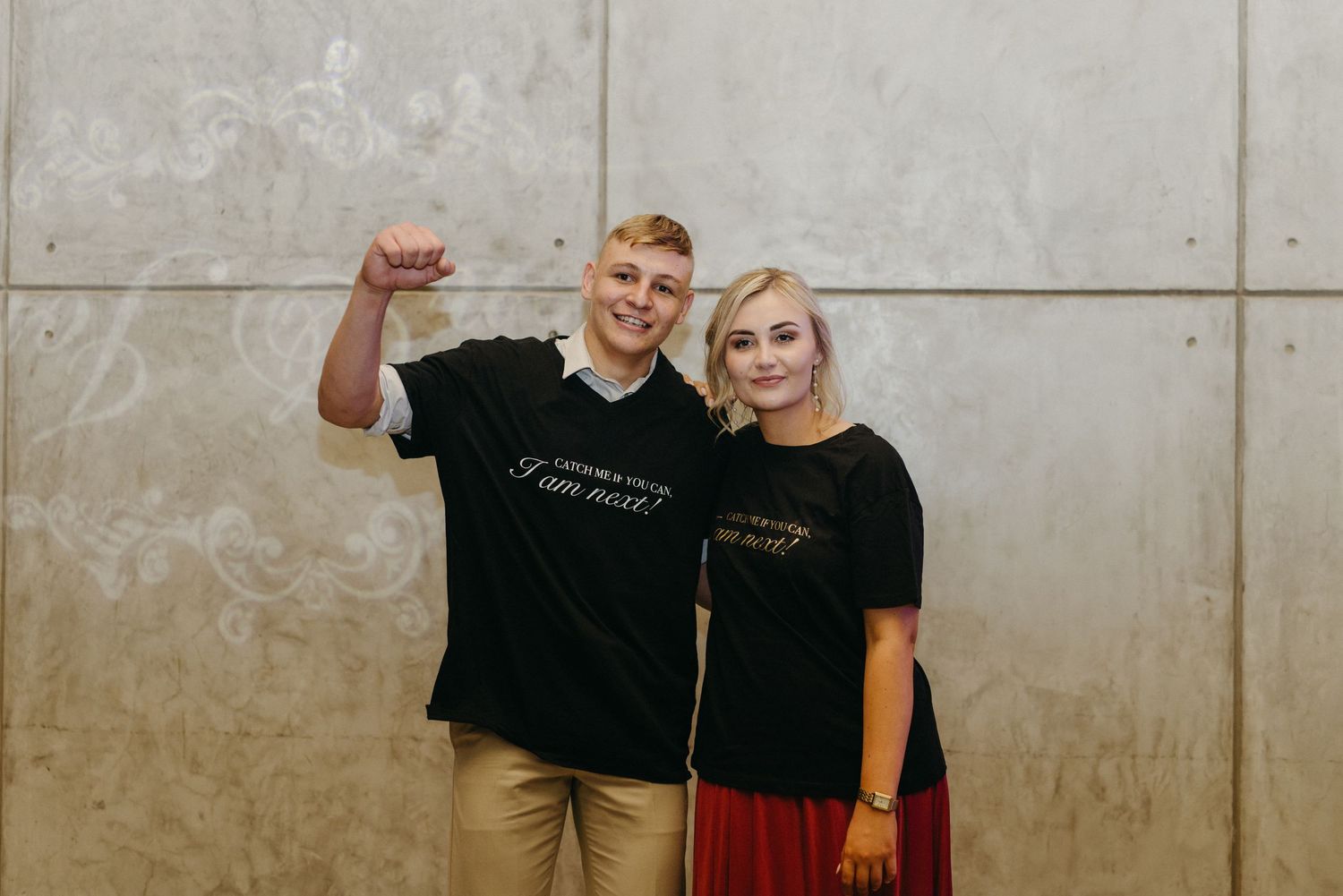 Two people wearing matching black t-shirts standing against a concrete wall smiling together.