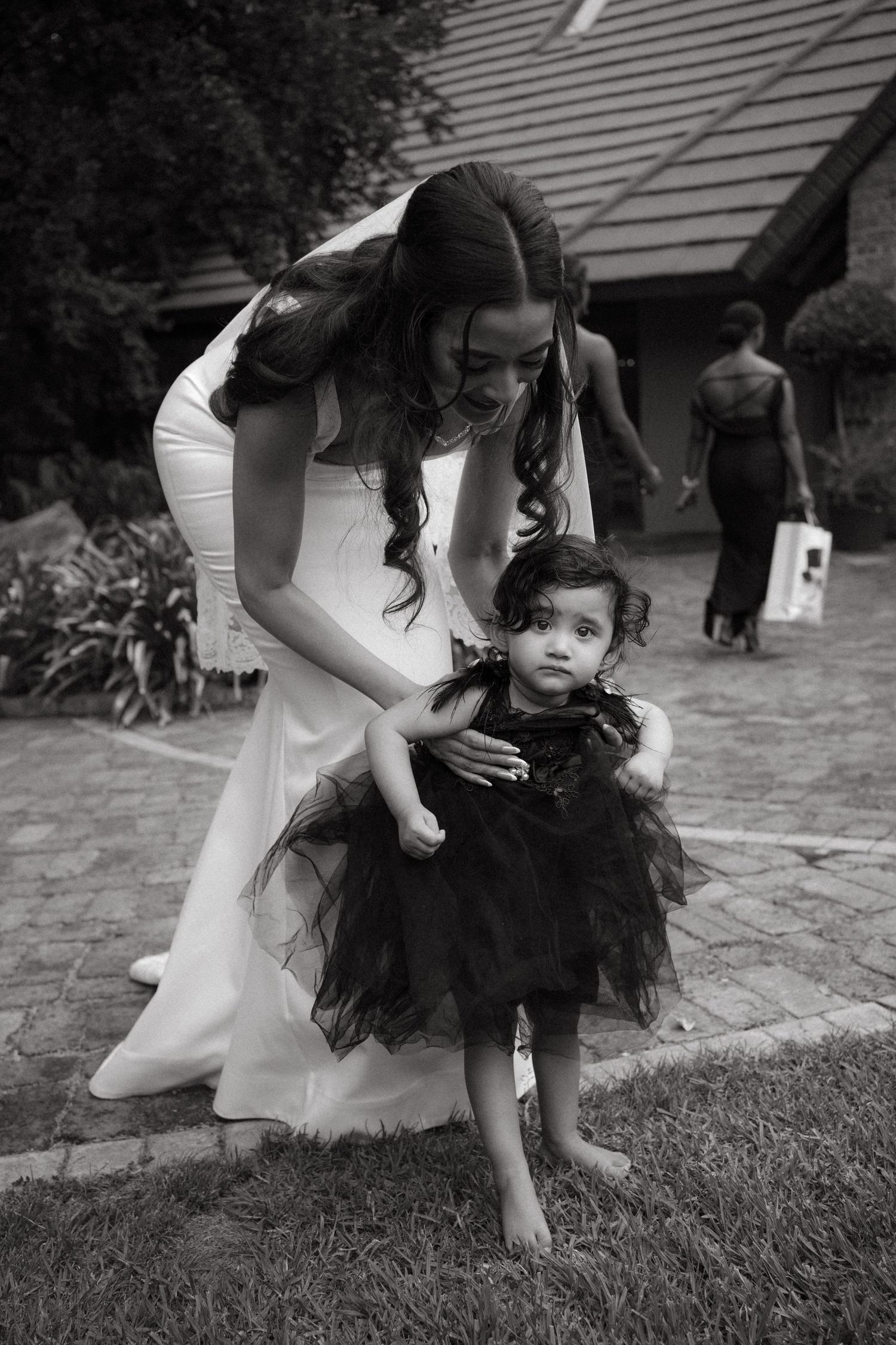 A woman in a white dress kneels beside a small child wearing a black tulle dress in this black and white photo.