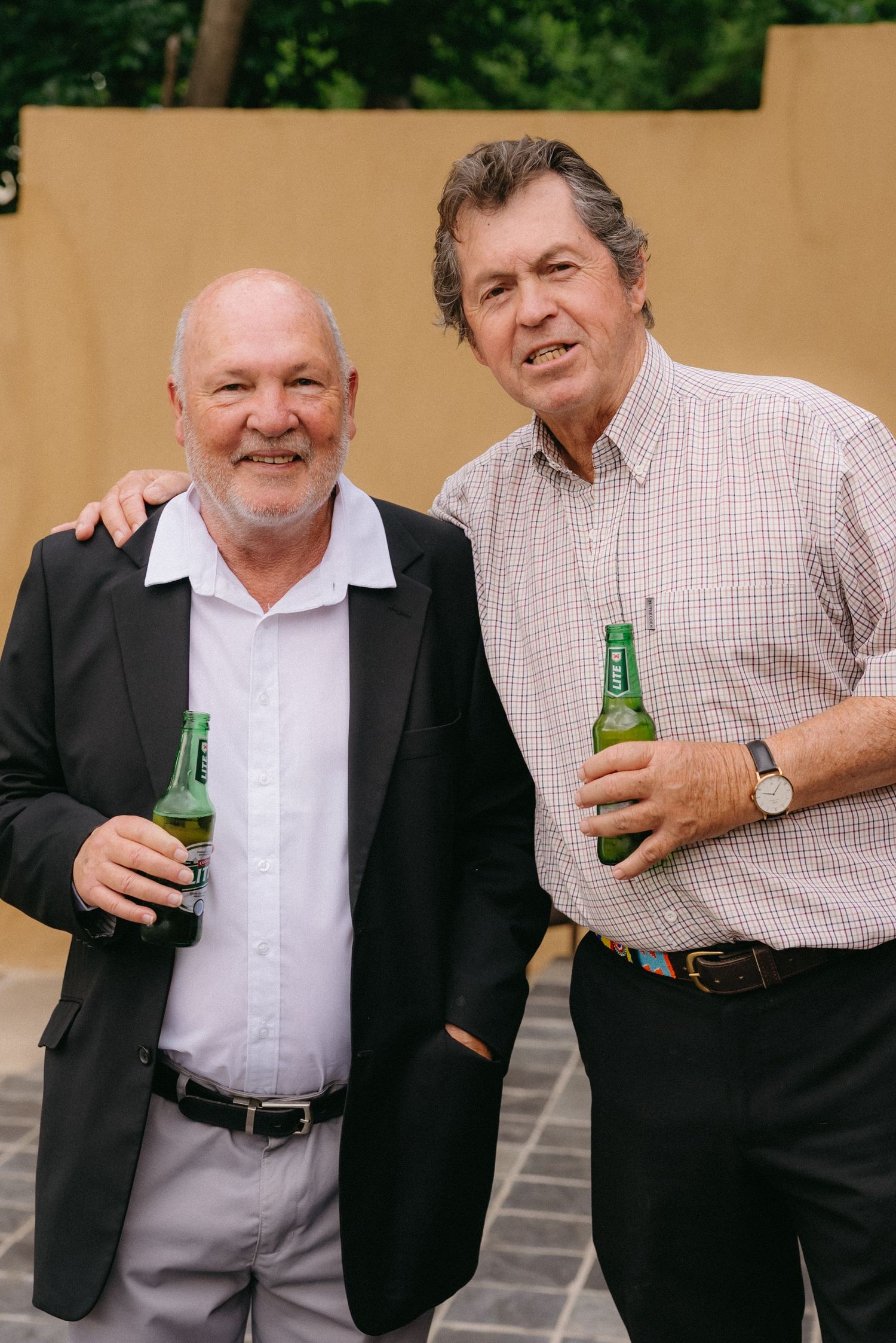 Two men in business attire share drinks and laughs at an outdoor gathering.