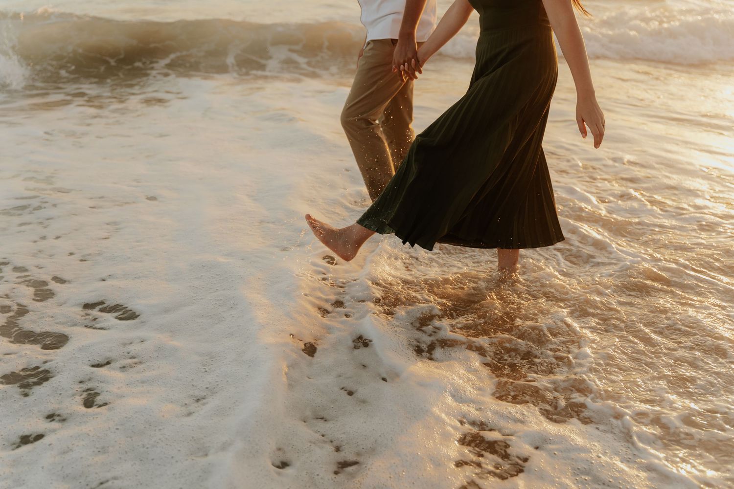 A person in a black dress splashes through shallow ocean water at sunset creating rippling reflections in the golden light.