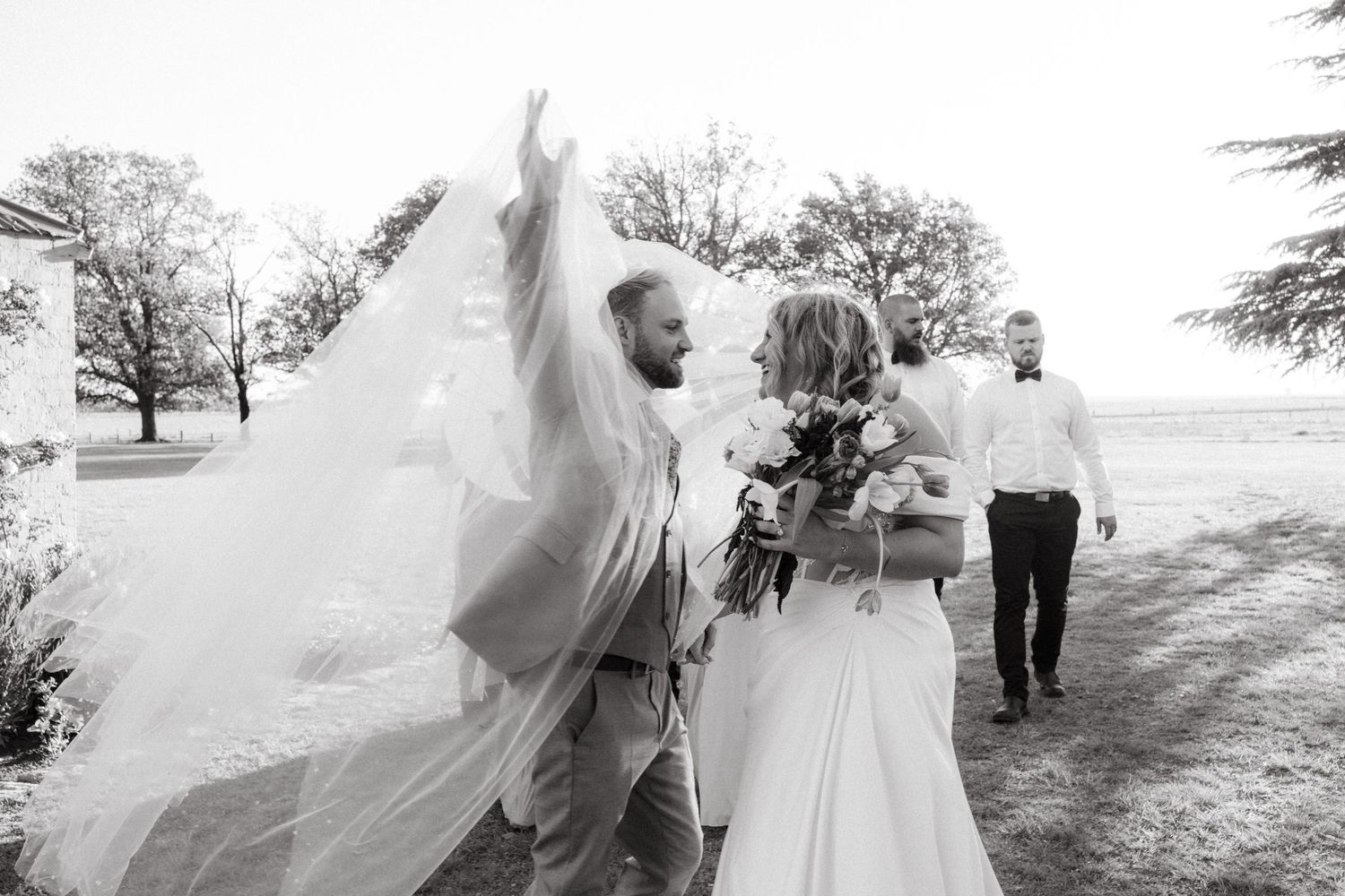 A joyful outdoor wedding scene with a flowing veil caught in the breeze.