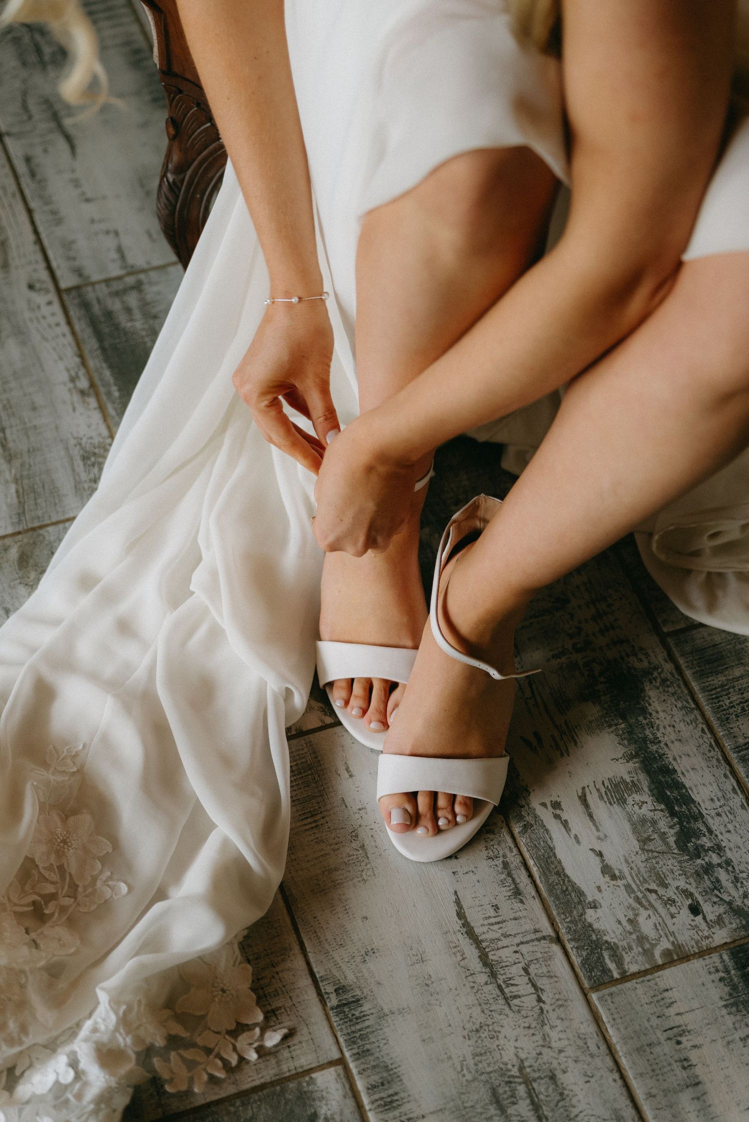 Close-up of bride putting on white wedding sandals on rustic wooden floor.