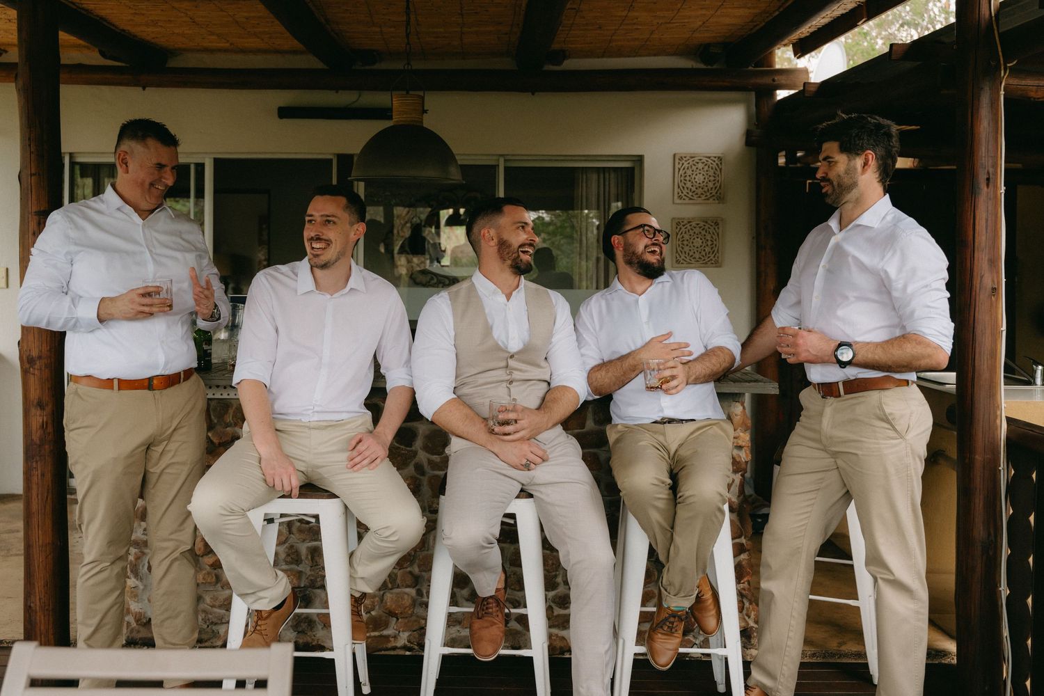 Five groomsmen in matching light colored attire share a candid laughing moment on a rustic porch.