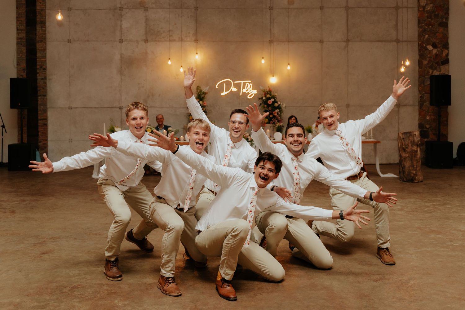 A wedding party in white attire poses playfully on the ground in a grand indoor venue with warm lighting.
