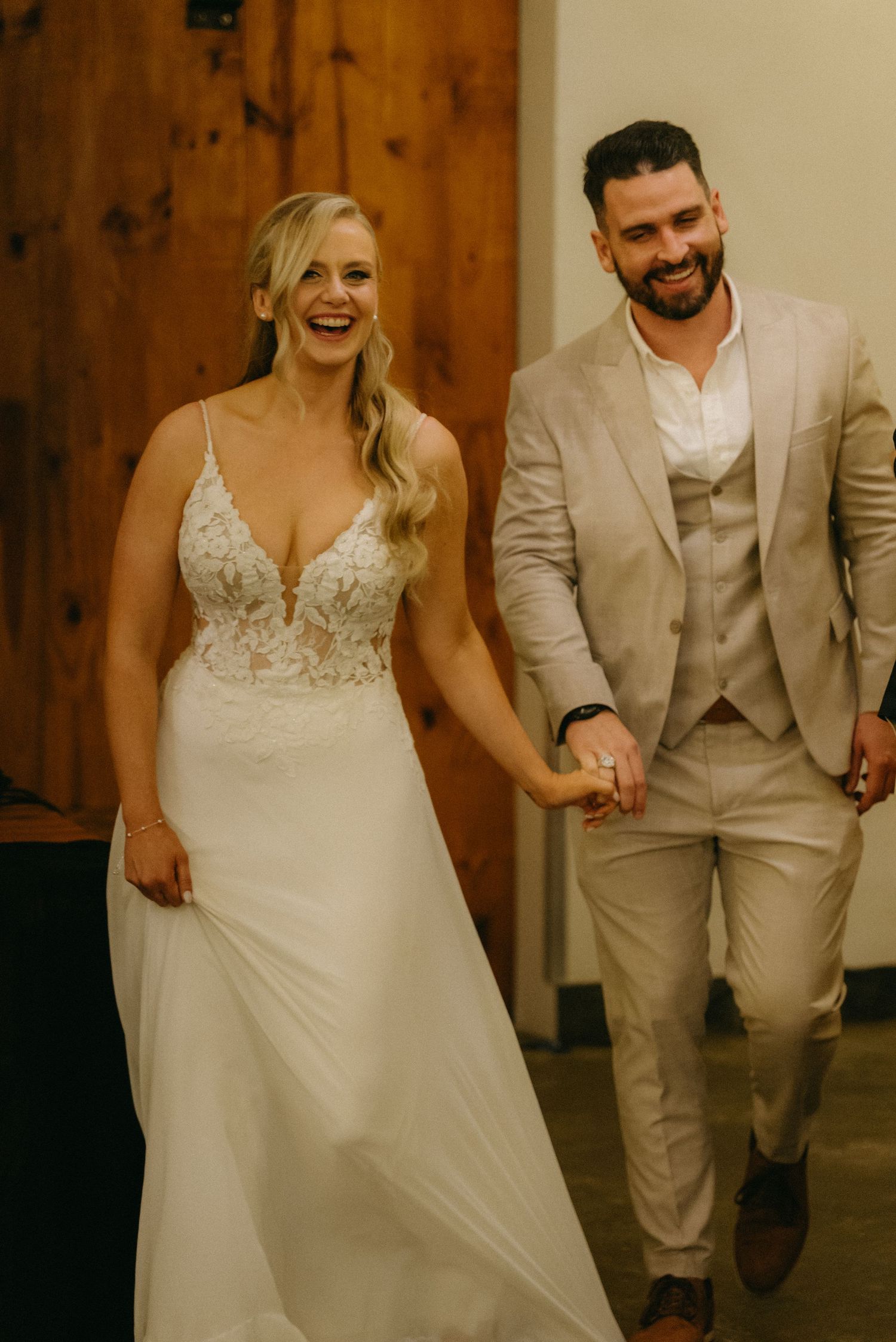 Bride and groom walk hand in hand through rustic indoor venue with wooden walls.