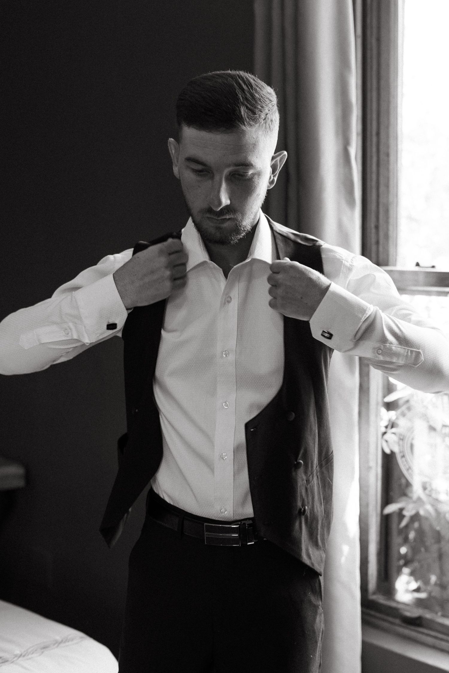 Black and white photo of person adjusting their suit and tie by window in dramatic lighting.