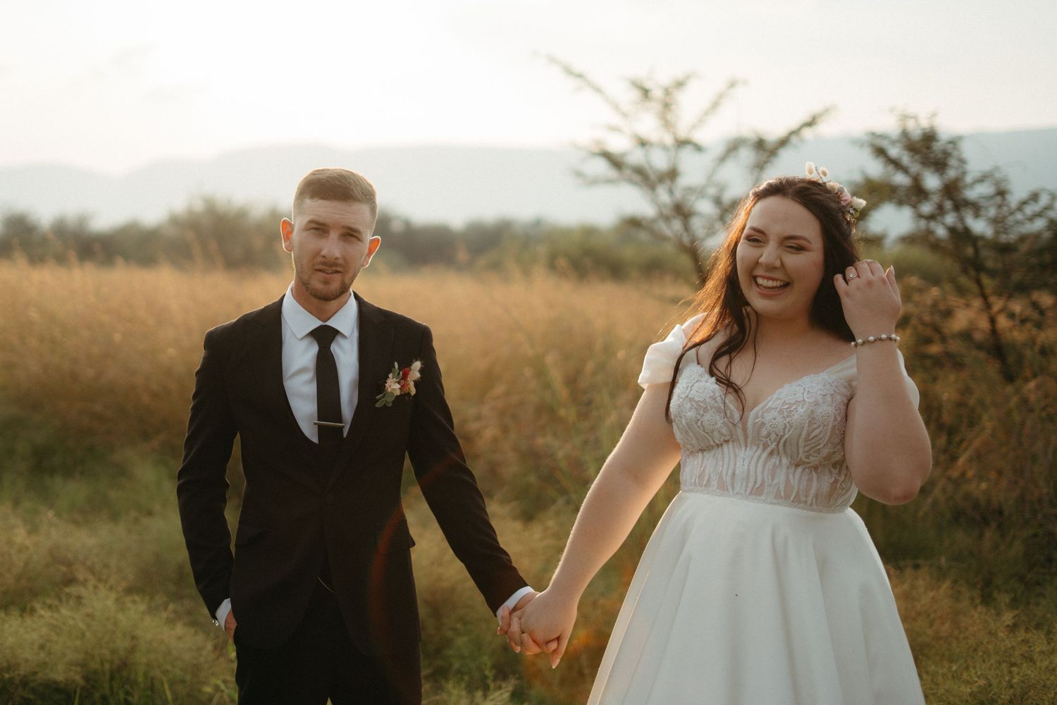 A couple poses together in formal wedding attire in a golden meadow at sunset.