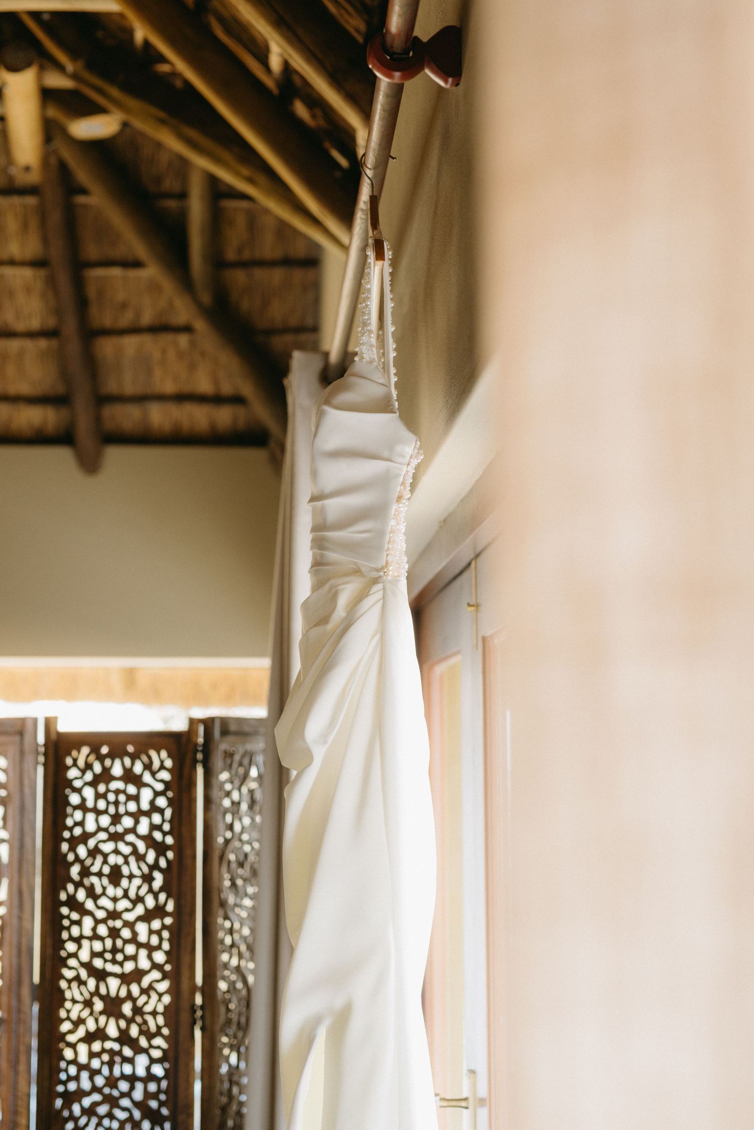 White wedding dress hanging in a rustic room with natural light streaming through decorative window screens.