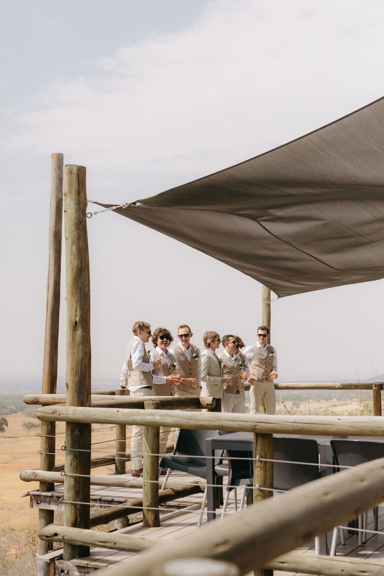 A group of people gather on a rustic wooden deck with canvas shade covers overlooking a scenic landscape.