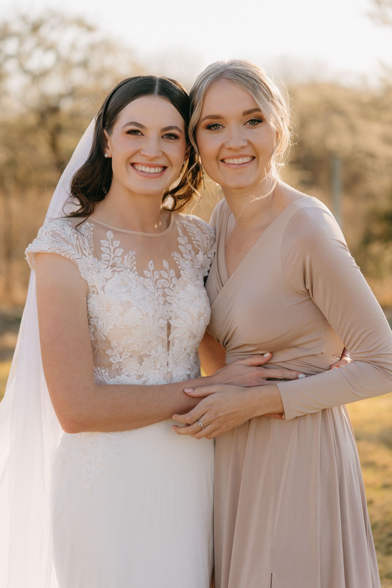 Two wedding attendants in white and beige dresses embrace while smiling outdoors in golden sunlight.