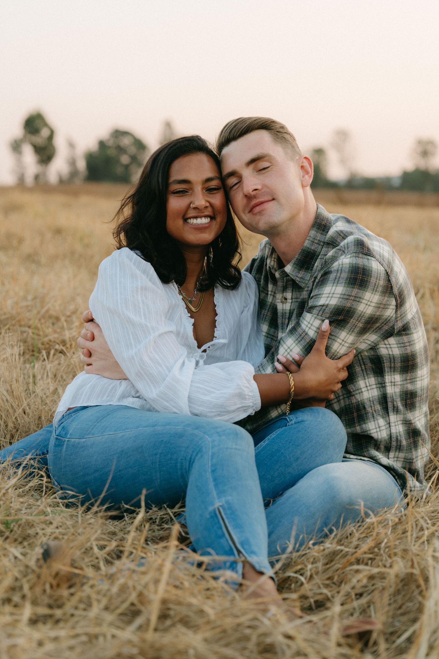 A couple laughs and embraces while sitting in a dry grass field at dusk.