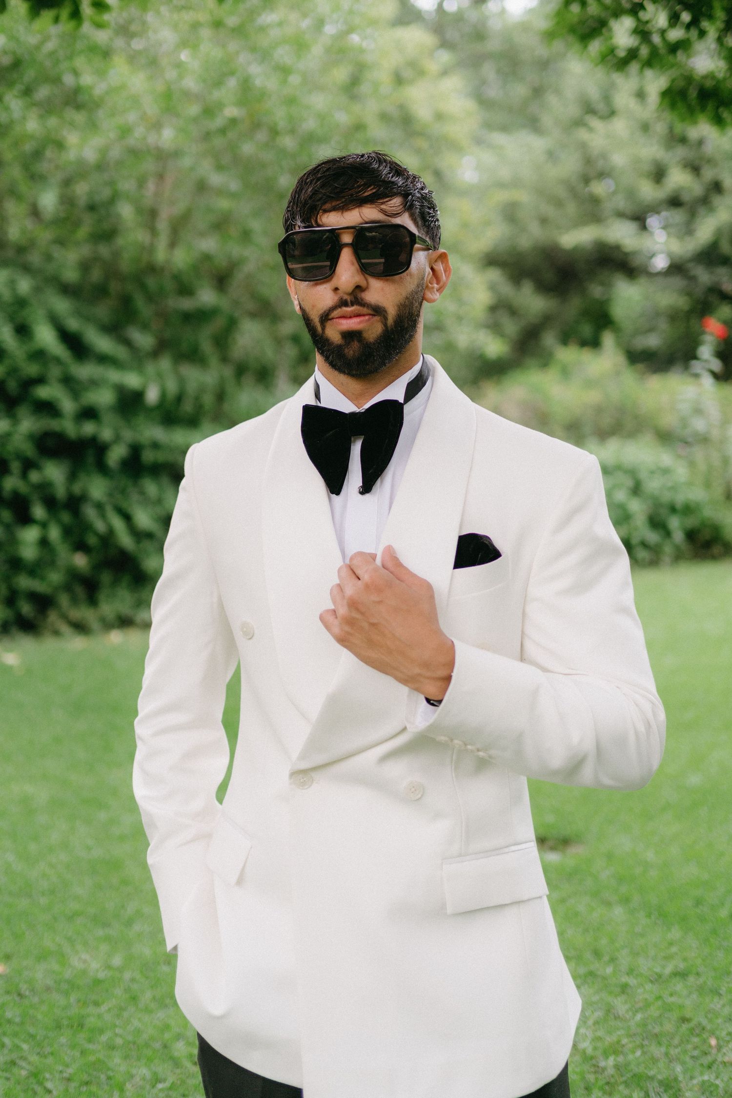 Groom in white tuxedo jacket and sunglasses poses in garden setting.