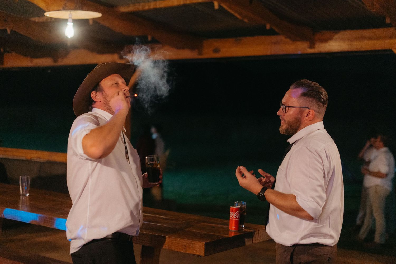 Two men engage in conversation on a patio at night, with one exhaling smoke while drinking from a can.