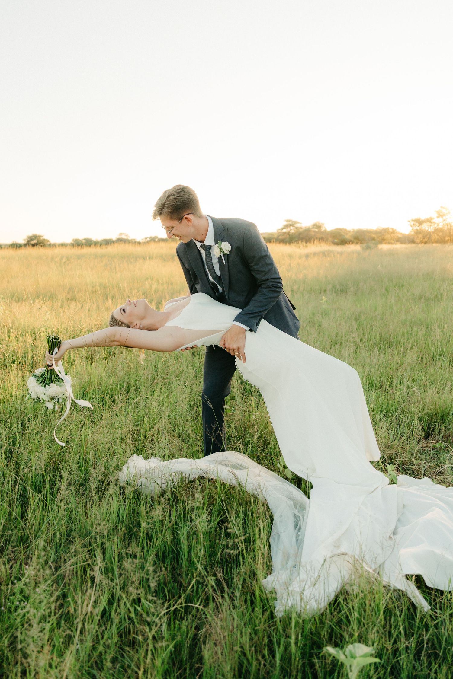 A romantic wedding couple dips dramatically in a sun-lit grassy field during their outdoor photo session.