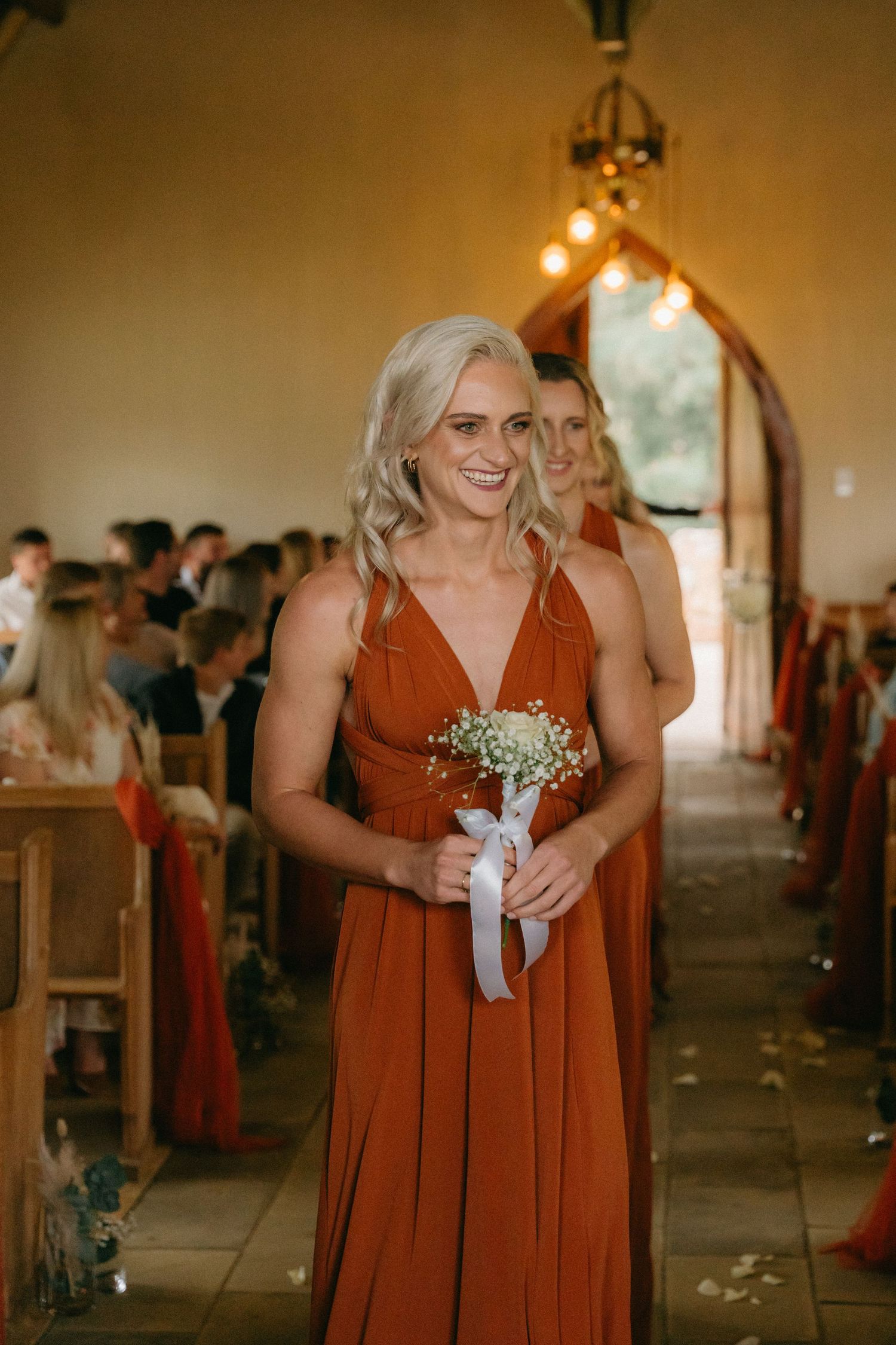 Bridesmaid in a flowing rust-colored dress holds a small bouquet of baby's breath in a church aisle.