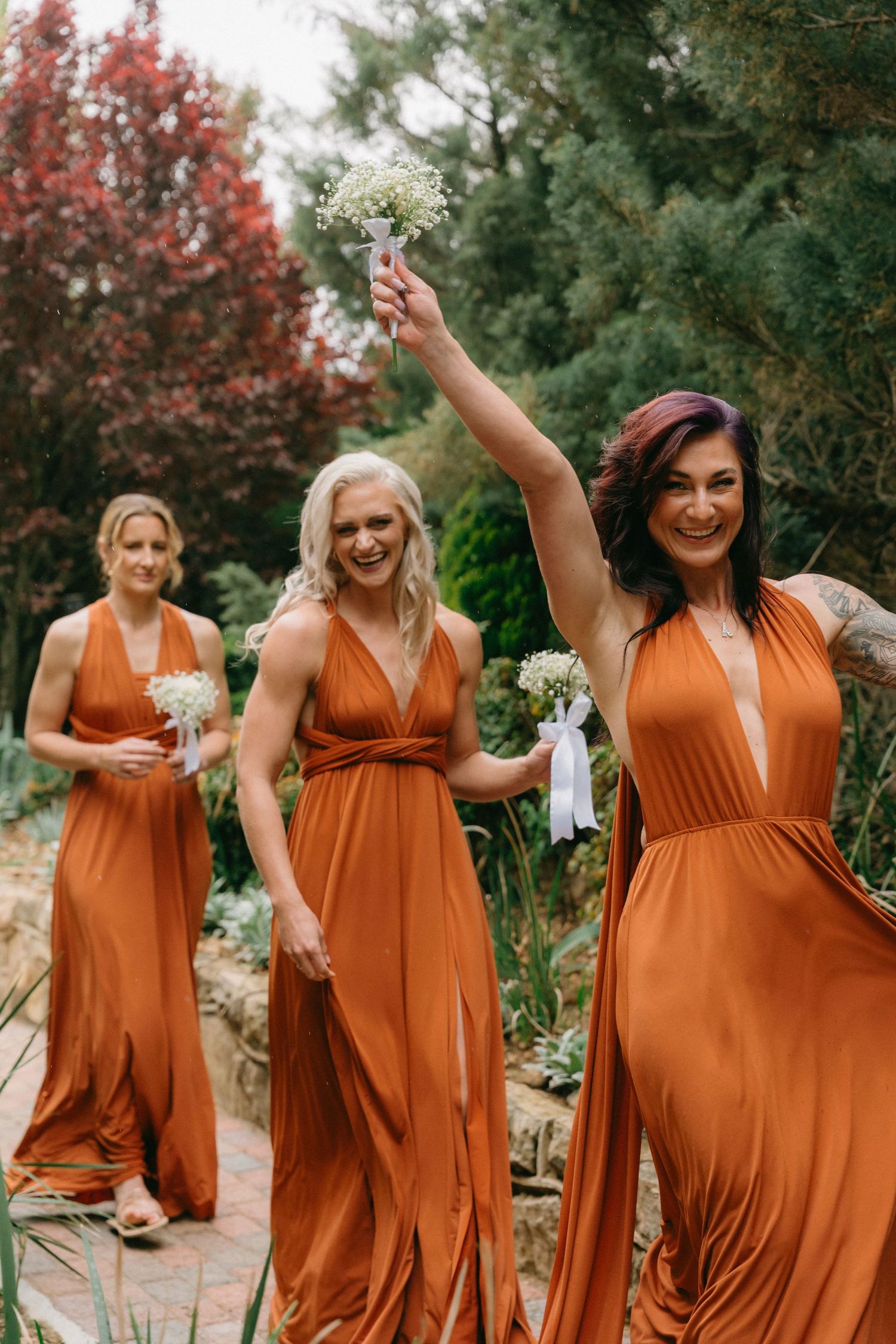 Bridesmaids in flowing rust-colored dresses dance and pose outdoors.
