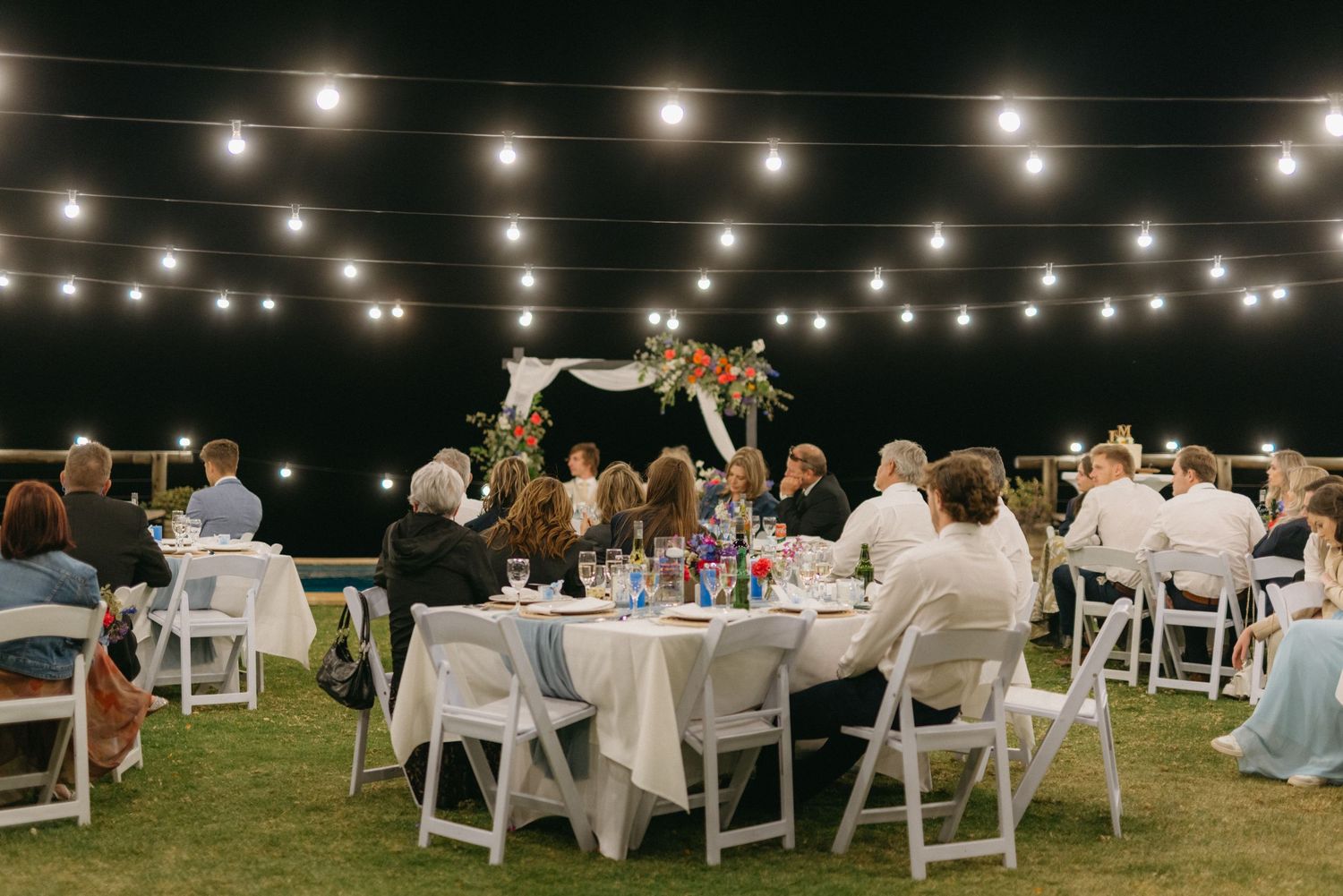 Outdoor wedding reception setup with string lights and white chairs under the night sky.