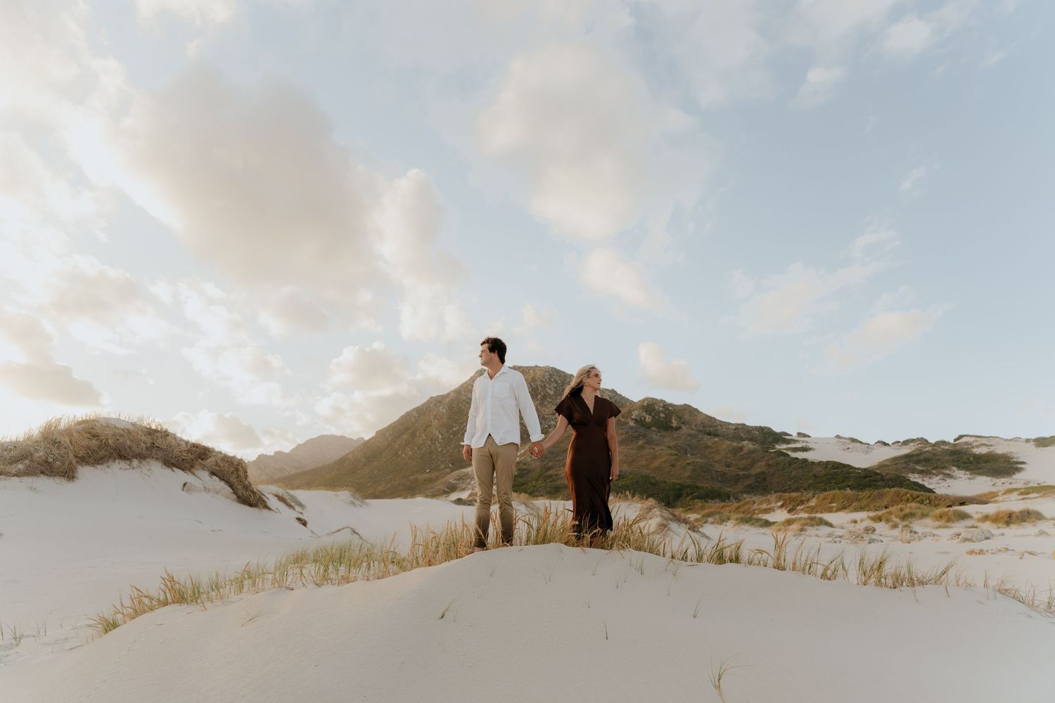 Couple holding hands on white sand dunes with green hills and cloudy sky in background.