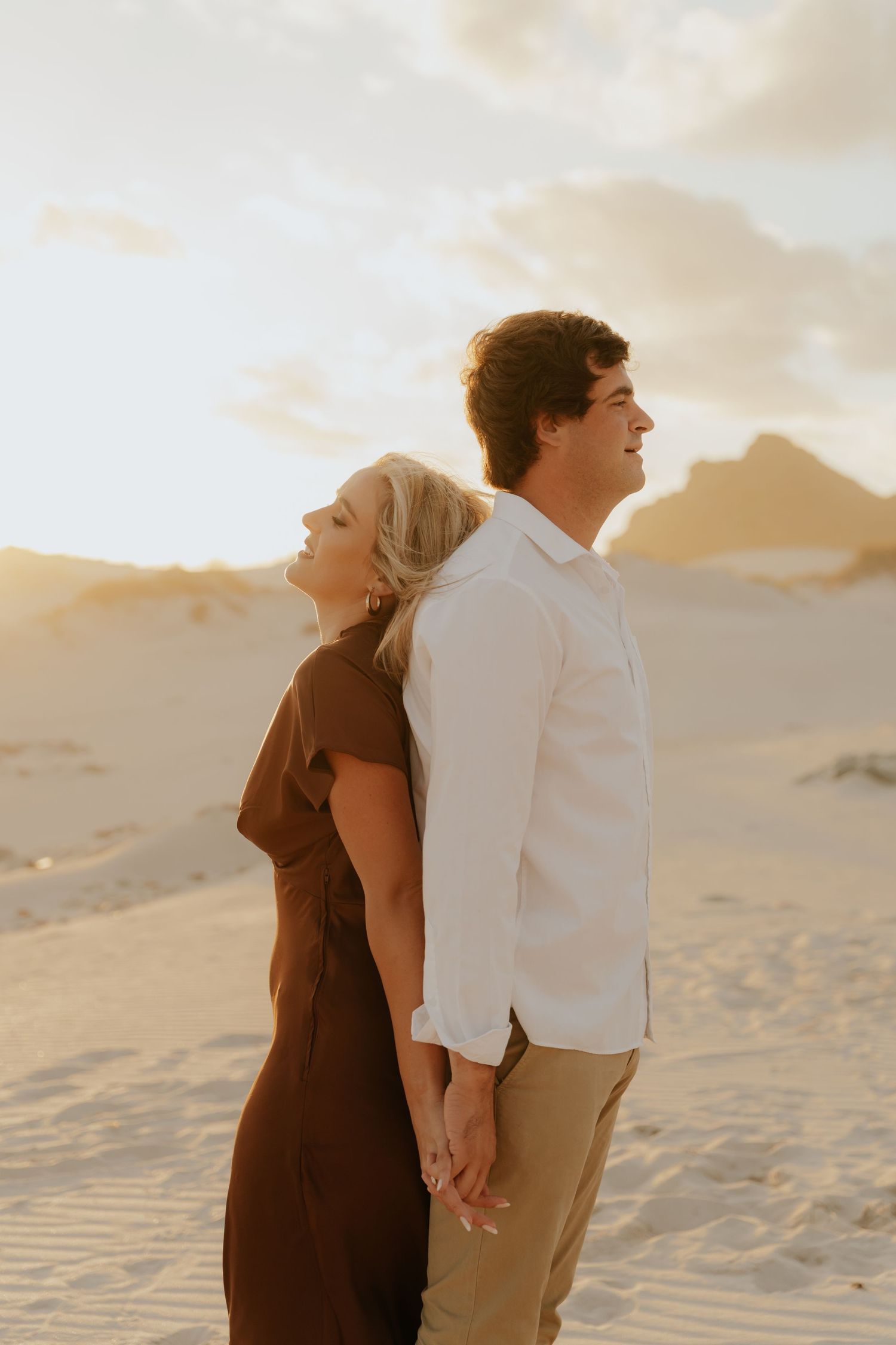 Couple holding hands back-to-back on sandy dunes at golden hour, woman in brown dress, man in white shirt.