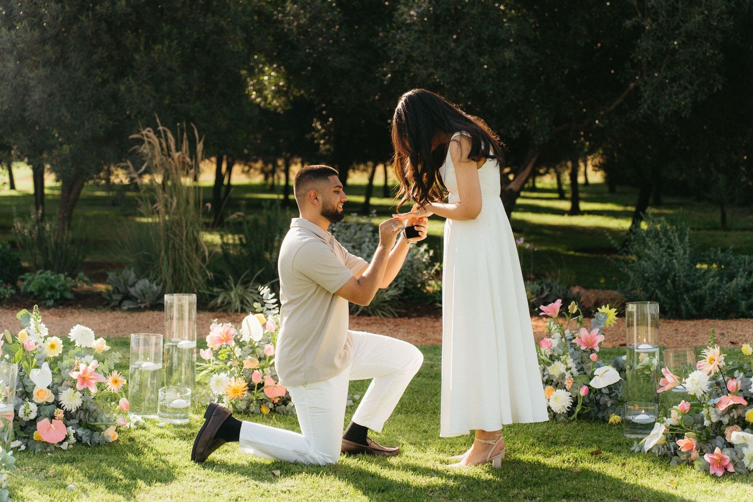 Man kneels on one knee proposing to woman in white dress at Ground the Venue in Muldersdrift, surrounded by floral arrangements.