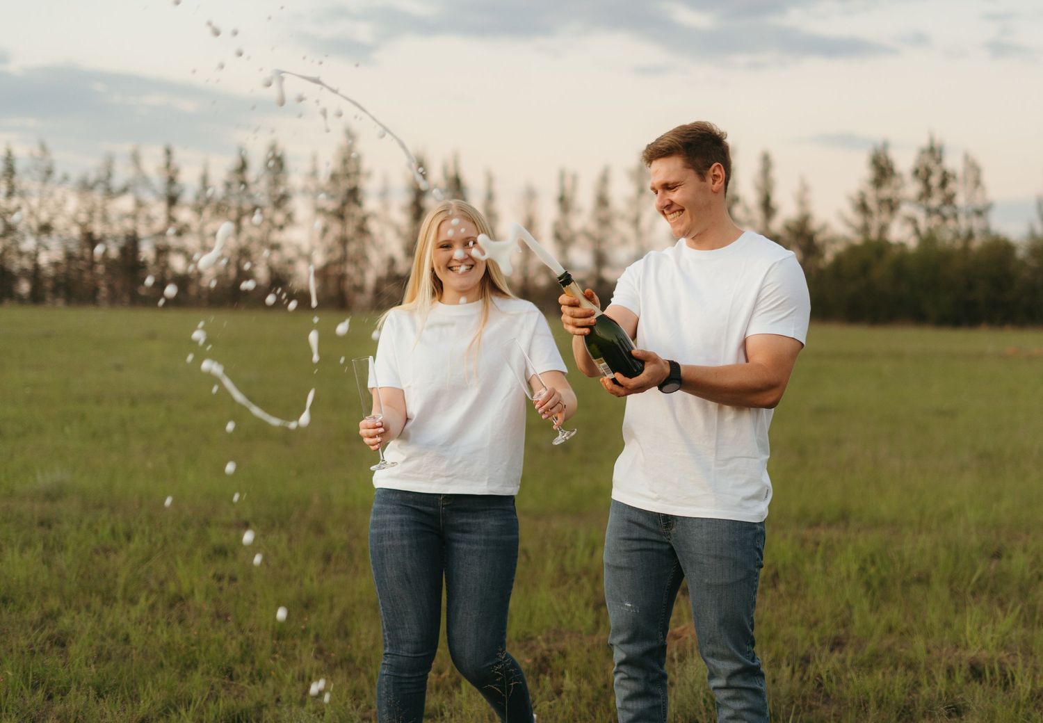 Happy couple in white shirts popping champagne in an open field.