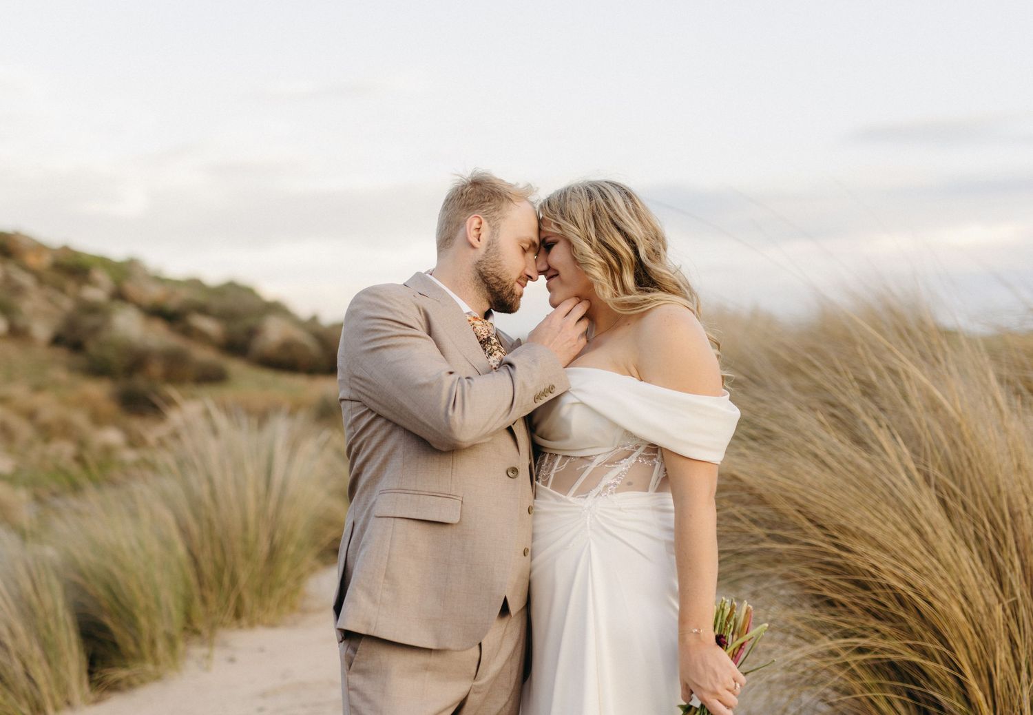 A romantic couple shares an intimate moment among coastal dunes with tall grass swaying in the breeze.