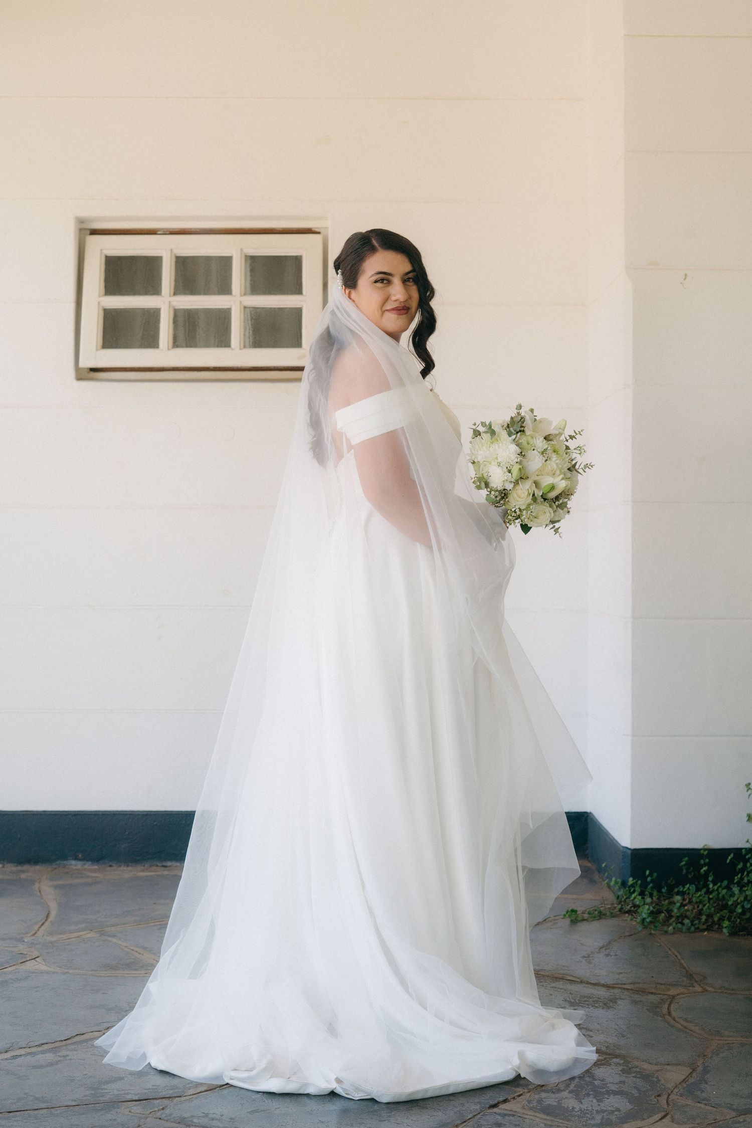 A bride in an elegant white off-shoulder wedding gown and veil poses with her bouquet against a light-colored wall.