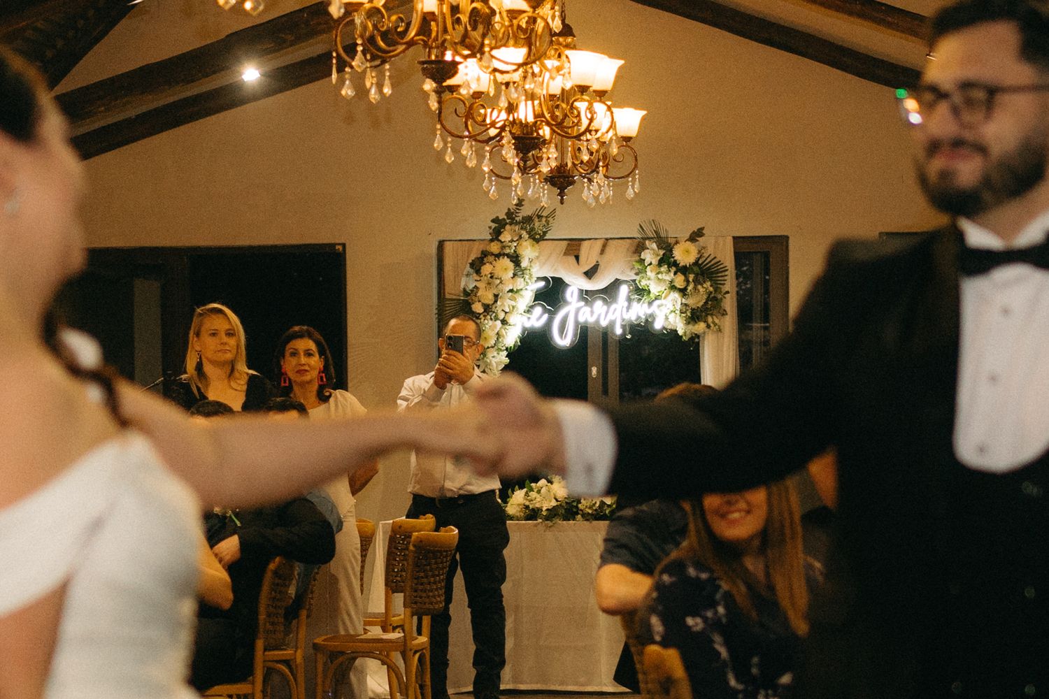 Couple dancing under an elegant chandelier during an evening reception while guests watch from the background.