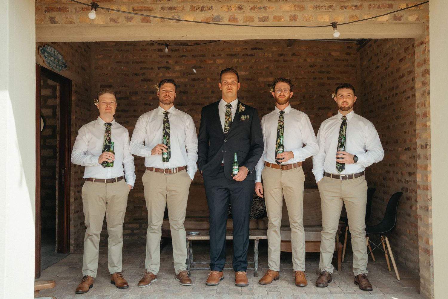 Wedding party in coordinated attire of khaki pants and white shirts posing against brick wall.