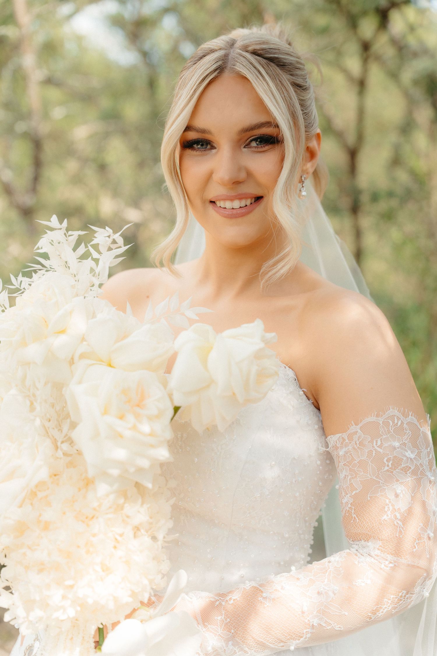 A radiant bride in a white lace gown holds a cream-colored bouquet against a natural outdoor backdrop.