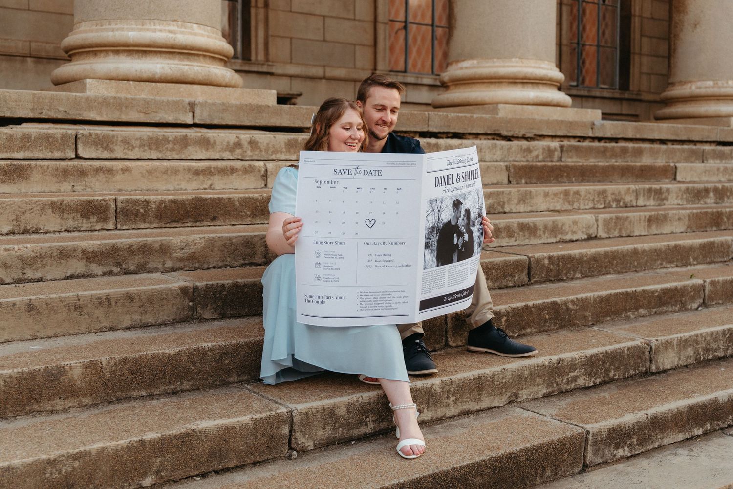 A couple sits on stone steps of a historic building while holding and reading a large newspaper together.