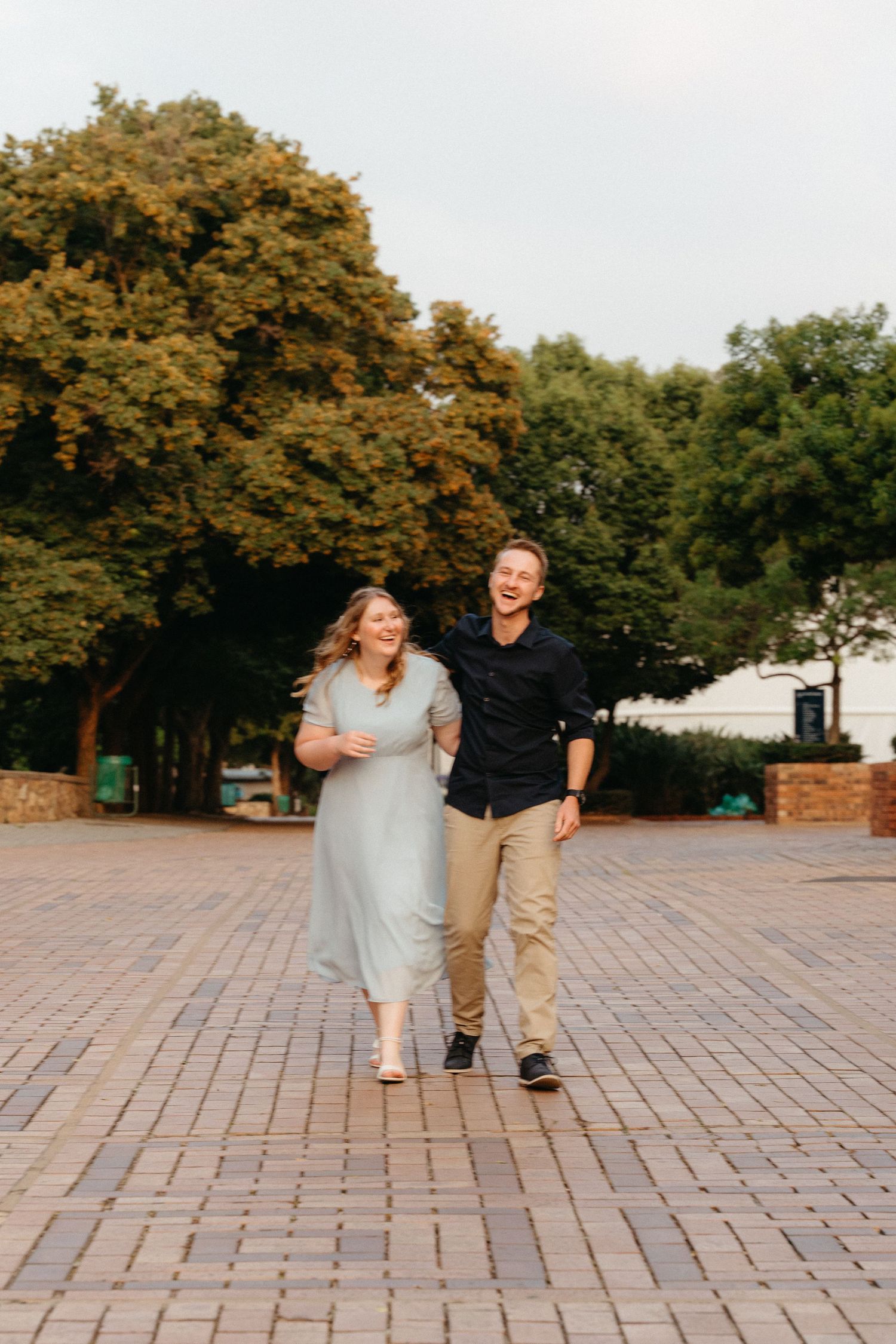 A couple shares a joyful moment walking together on brick pavement with autumn trees in the background.